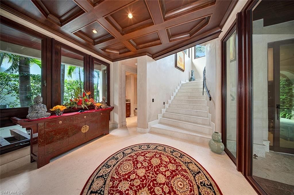 This interior shot showcases an elegant entryway featuring a coffered wood ceiling, a red oriental rug, and a dark wood console table adorned with floral arrangements and a Buddha statue. A staircase with white steps and a black railing leads to the upper level, while large windows provide natural light and views of the exterior greenery. The overall impression is one of sophisticated luxury and tranquility.