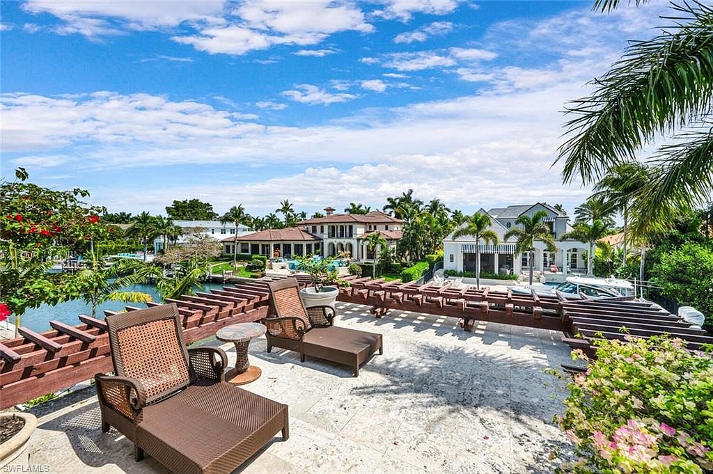 This image showcases a luxurious patio or deck area with comfortable lounge chairs and a small table, suggesting an ideal space for relaxation and outdoor entertainment. The patio features a pergola-style structure providing partial shade, and the backdrop includes waterfront views, lush landscaping, and upscale residential architecture. The overall impression is one of high-end living and serene outdoor enjoyment.