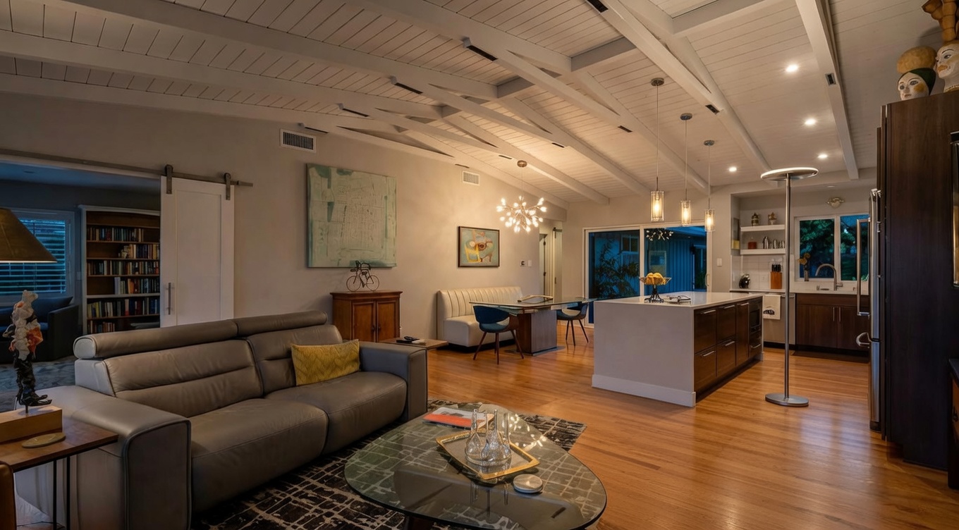 This open-concept living space exudes a mid-century modern aesthetic, featuring vaulted ceilings with exposed white beams and warm hardwood flooring. The room seamlessly integrates a comfortable lounge area with a grey sofa and a mustard accent chair, a dedicated dining corner with a booth-style setup, and a well-appointed kitchen in the background. Abundant recessed and pendant lighting, combined with neutral wall tones, create a bright, inviting atmosphere perfect for contemporary family living.