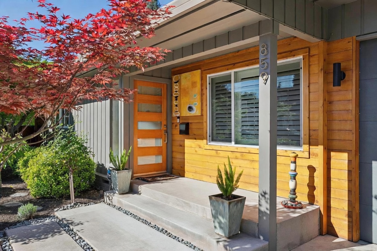 This inviting entryway features a modern wood-paneled facade, a sleek glass-paneled front door, and a concrete porch adorned with potted plants. A vibrant Japanese maple tree adds a pop of red to the left, while a unique decorative sculpture stands near the porch steps. The overall aesthetic is contemporary and warm, creating a welcoming curb appeal.