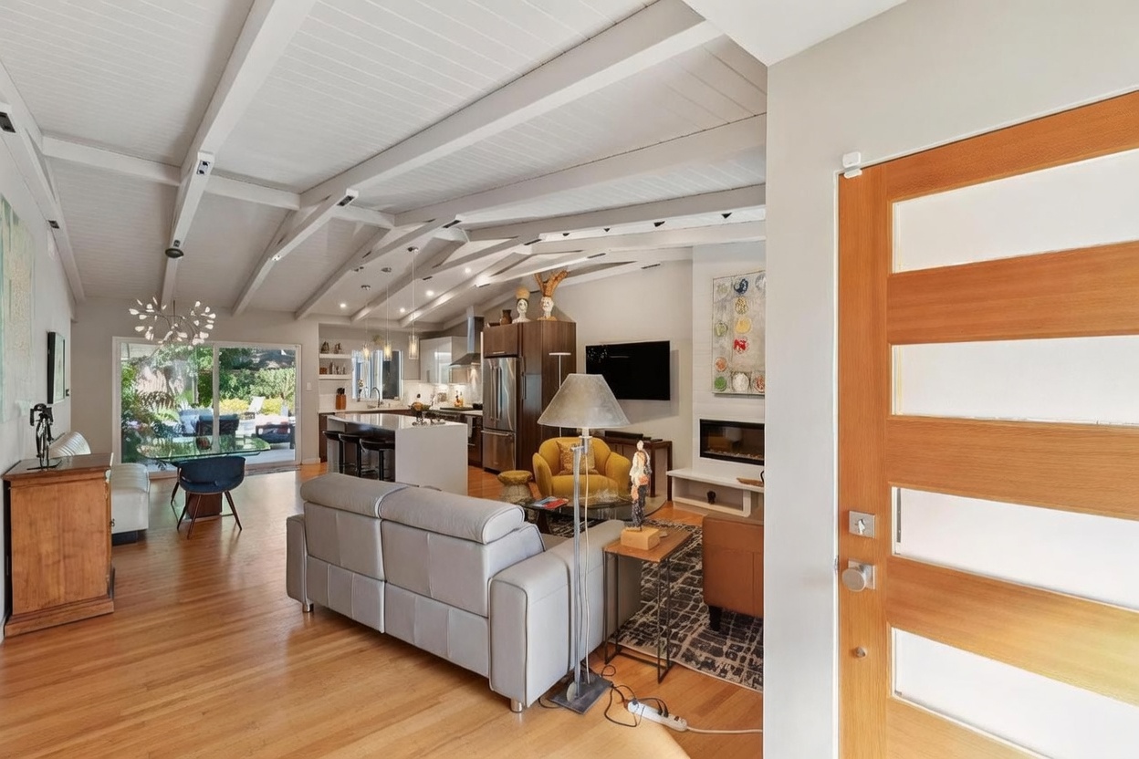 This open-concept living space features a vaulted ceiling with exposed white beams and warm hardwood flooring, creating an airy and inviting atmosphere. The room seamlessly integrates a cozy seating area with a fireplace, a modern kitchen with a central island, and a dining space that opens to the outdoors through large sliding glass doors. The perspective is from the entryway, looking into the heart of the home, highlighting the spacious and cohesive design.