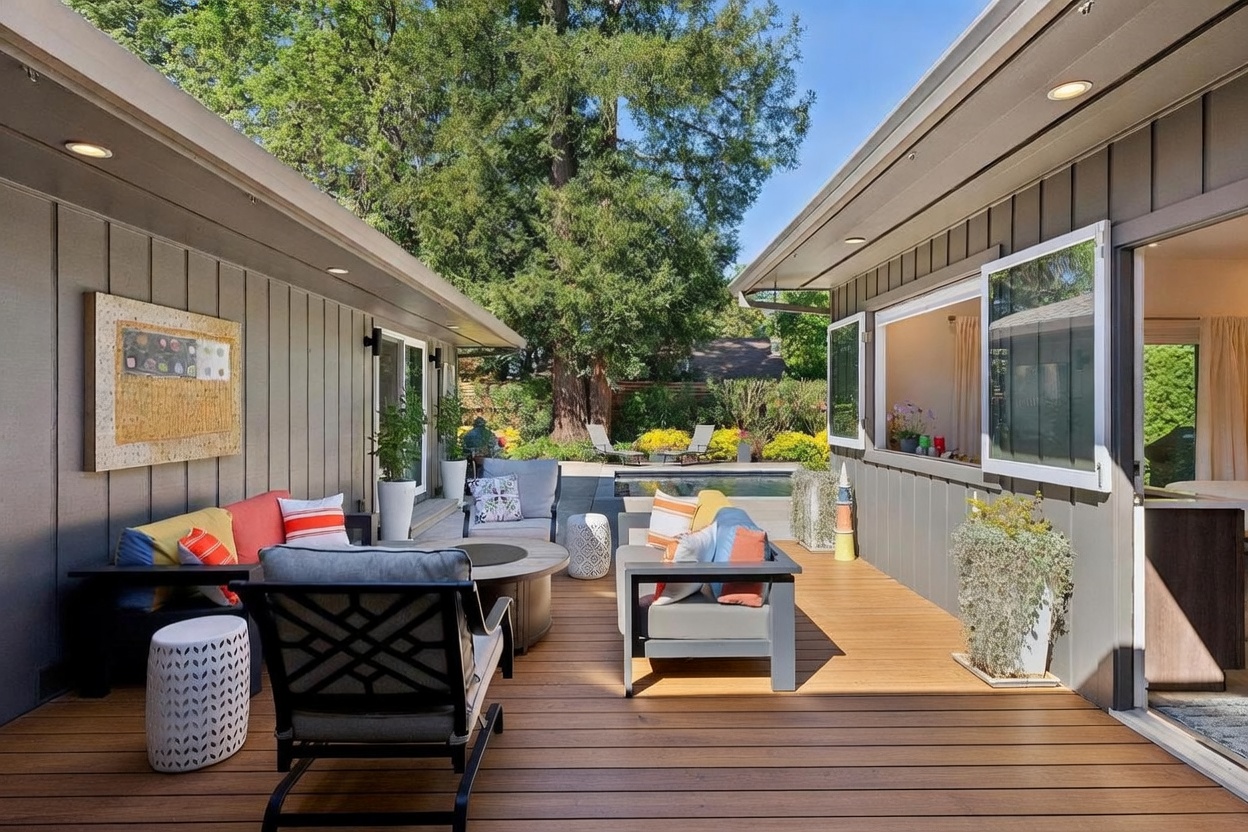 This inviting outdoor living space features a spacious wooden deck furnished with comfortable modern seating, colorful accent pillows, and a central fire pit. The area is framed by two wings of the house, creating a cozy courtyard feel that opens up to a swimming pool and lush backyard greenery in the distance. The scene is bathed in bright, natural sunlight, highlighting the clean lines of the architecture and the relaxed, resort-style atmosphere.
