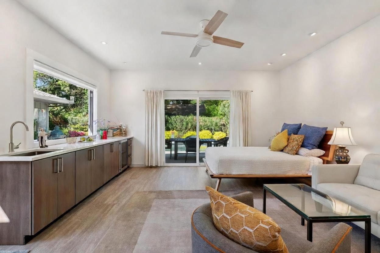 This versatile studio-style bedroom features a modern kitchenette with dark wood cabinetry and a sleek countertop, seamlessly integrated into the living space. A comfortable bed with blue and yellow accents sits near a large sliding glass door that opens to a lush, green backyard patio. The room is bright and airy, characterized by light wood-look flooring, a contemporary ceiling fan, and a cozy seating area with a glass-top coffee table.
