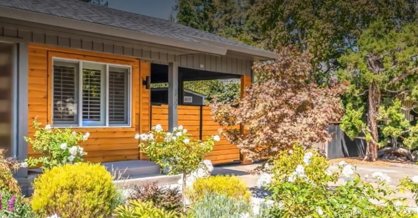 This image showcases the appealing front exterior of a home, featuring warm wood-paneled siding and a covered entryway. The facade is accented by large, multi-pane windows and lush landscaping with flowering shrubs, creating an inviting and well-maintained curb appeal.
