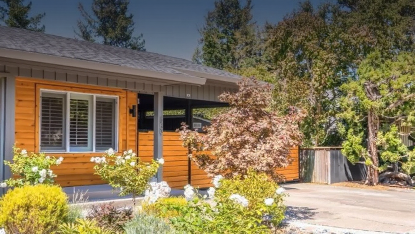 This single-story home features a warm, horizontal wood-paneled exterior siding accented by gray trim and a neutral-toned roof. A prominent covered carport area and a well-maintained front garden with blooming shrubs and greenery create an inviting curb appeal. The overall aesthetic is a charming blend of contemporary design with a natural, rustic touch.