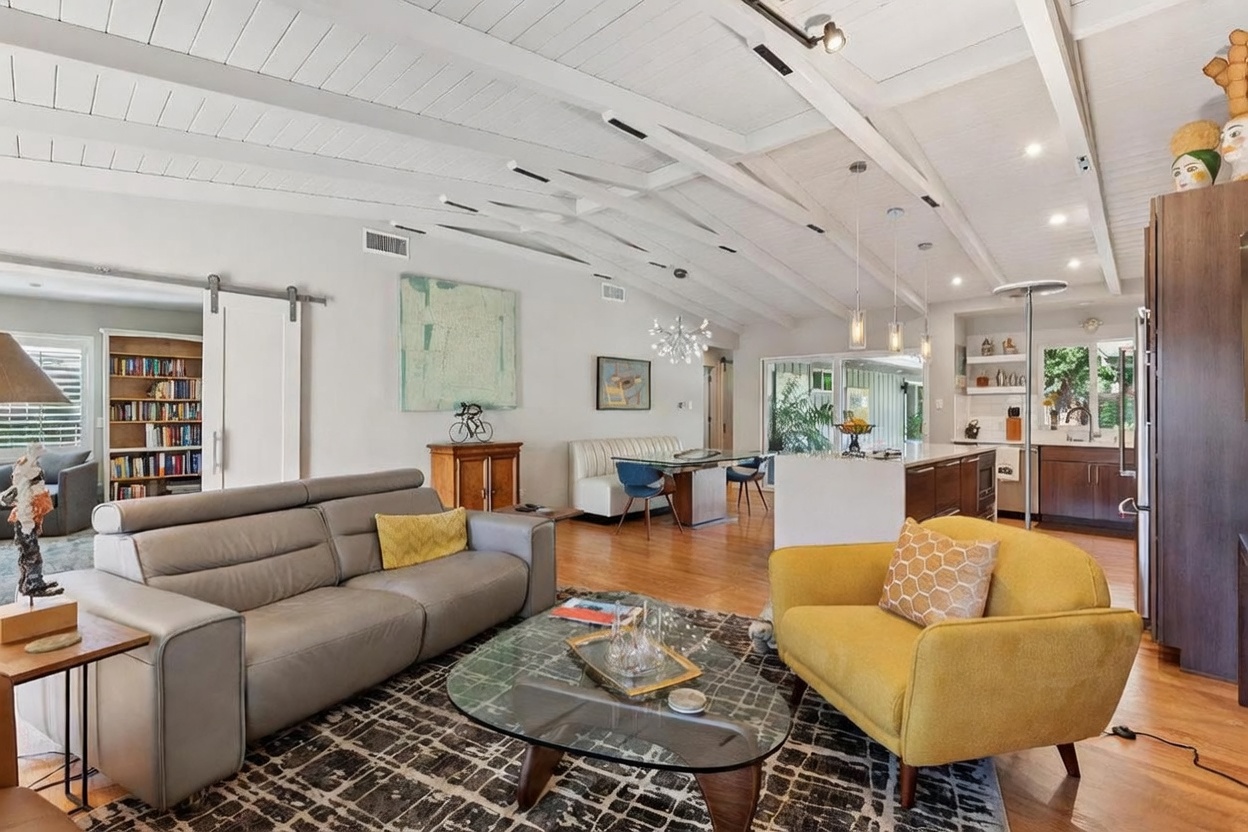 This open-concept living space features a mid-century modern aesthetic with vaulted white-beamed ceilings and warm hardwood flooring. The room is anchored by a grey leather sofa and a vibrant yellow armchair, complemented by a geometric area rug and a unique glass-topped coffee table. The space flows seamlessly into a dining area and kitchen, creating an airy, inviting atmosphere perfect for entertaining.