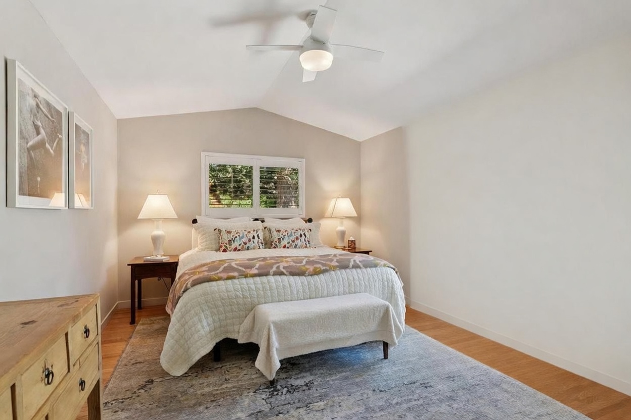 This bright and airy primary bedroom features a vaulted ceiling, a plush queen-sized bed with decorative pillows, and a matching bench at the foot. Large sliding glass doors provide a seamless transition to the outdoor pool area, flooding the room with natural light. The neutral color palette, warm wood flooring, and elegant bedside lamps create a serene and inviting atmosphere.