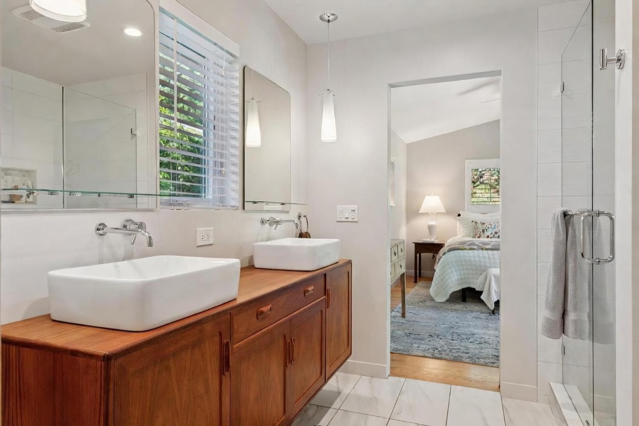 This modern primary bathroom features a sleek double vanity with white vessel sinks atop a warm-toned wooden cabinet. The space is bright and airy, with a glass-enclosed shower and a direct view into an adjacent bedroom, creating a seamless flow between rooms. The clean lines, marble-look tile flooring, and minimalist fixtures contribute to a sophisticated, contemporary aesthetic.
