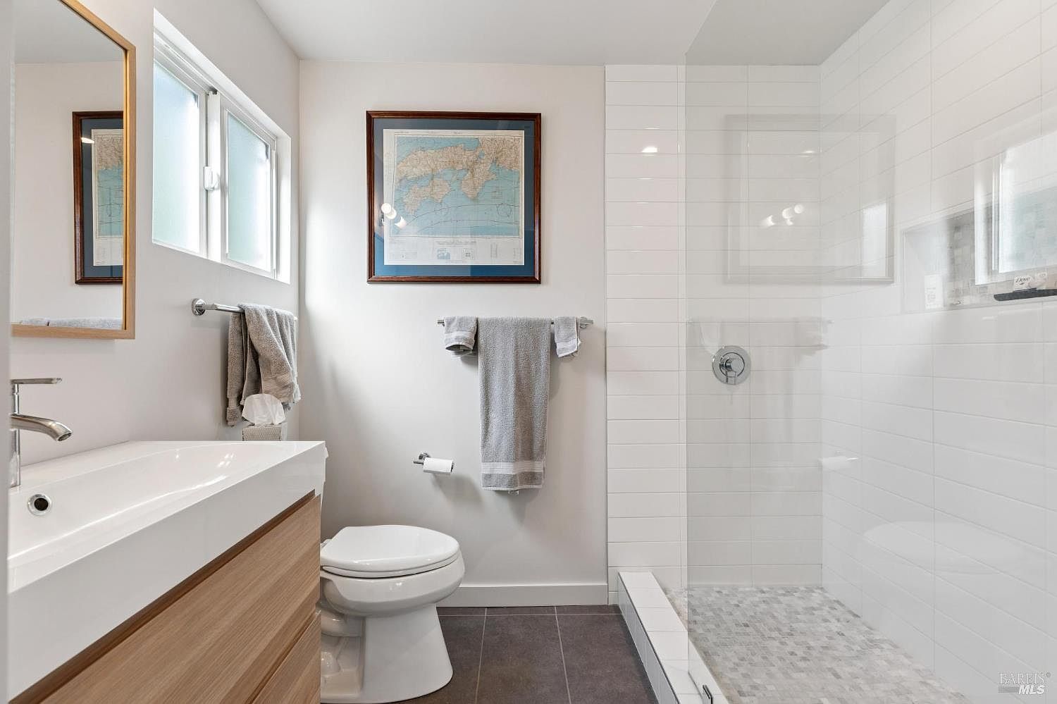 This modern, minimalist bathroom features a sleek floating vanity with a wood-grain finish and a white countertop, paired with a clean, white-tiled walk-in shower. The space is accented by dark grey floor tiles, a framed map on the wall, and a large mirror, creating a bright and contemporary aesthetic. The perspective is a straight-on shot that highlights the efficient layout and clean lines of the room.