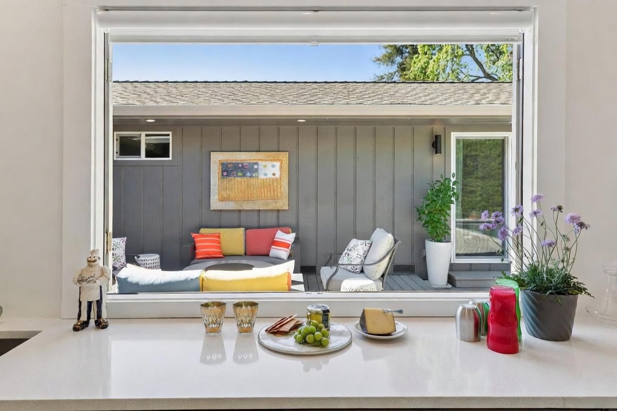 This perspective captures a bright kitchen pass-through window looking out onto a cozy, modern outdoor patio area. The foreground features a clean white countertop with a small snack arrangement, while the background showcases a vibrant outdoor seating area with colorful cushions and a decorative wall art piece. The scene creates a seamless indoor-outdoor living experience, emphasizing a relaxed and inviting atmosphere.