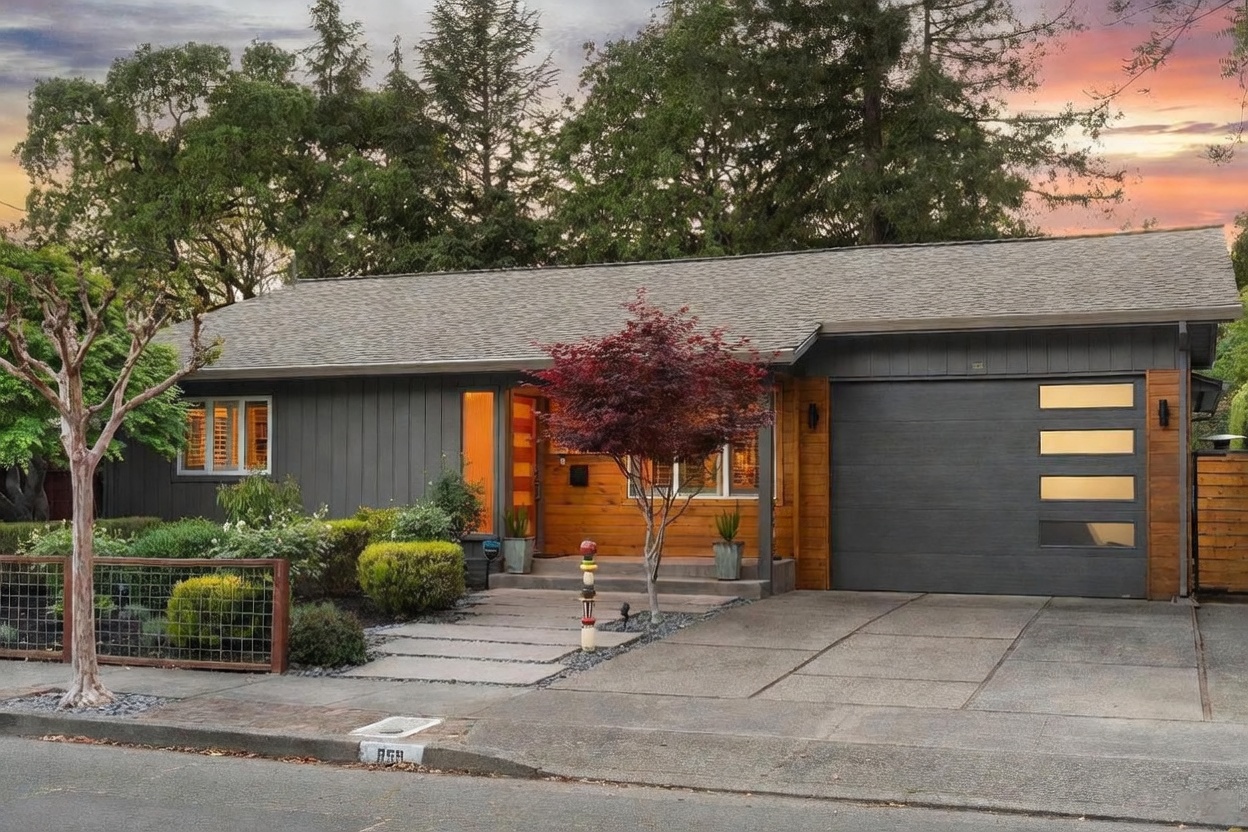 This charming single-story home features a modern aesthetic with a striking combination of dark vertical siding and warm wood paneling. The front facade includes a prominent garage with horizontal windows, a welcoming entryway with a Japanese maple tree, and a clean concrete walkway leading to the front door. The scene is captured at dusk, with the warm interior lighting creating an inviting and cozy atmosphere against the backdrop of a sunset sky.