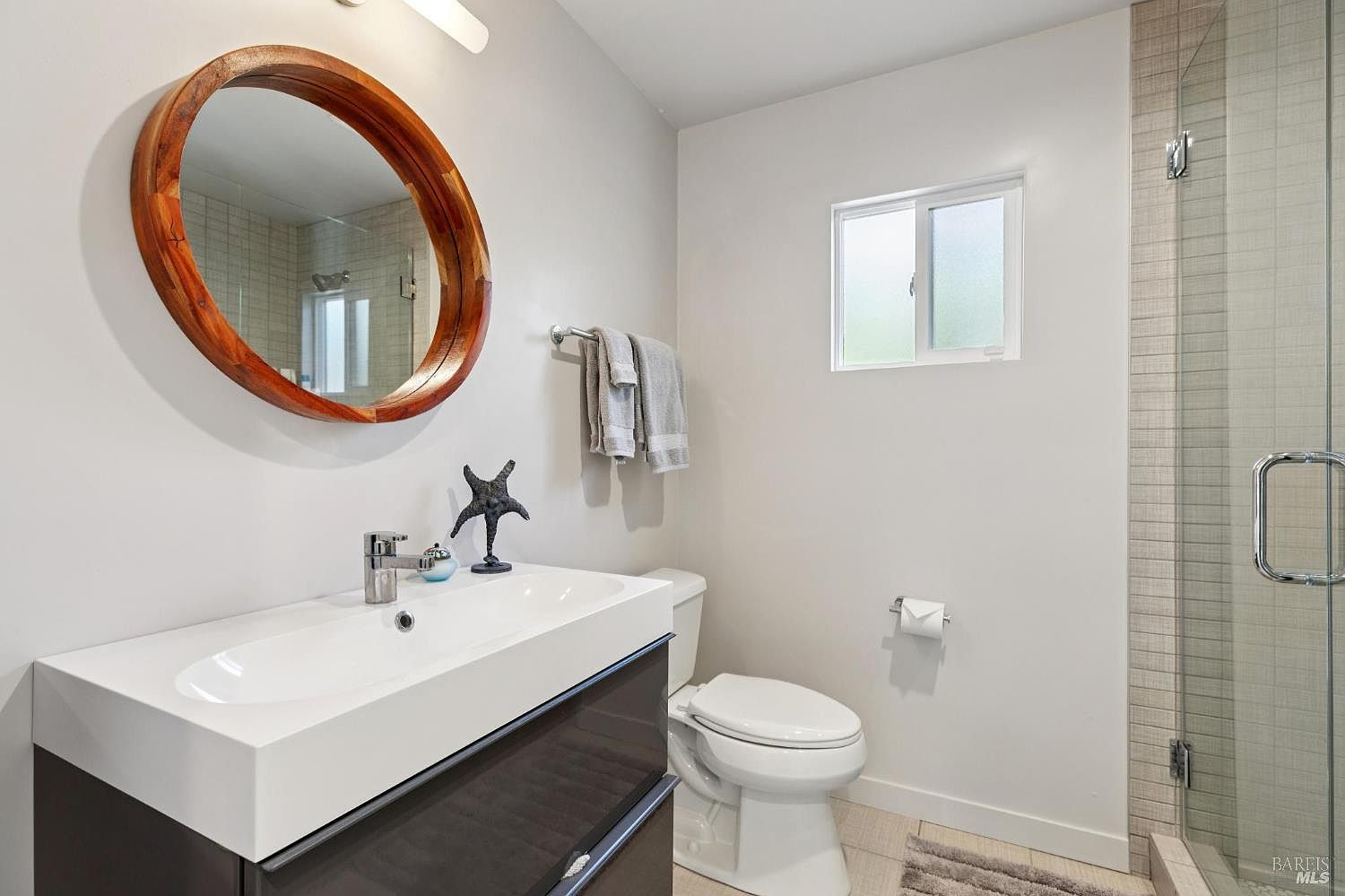 This modern bathroom features a sleek, dark-toned vanity with a white rectangular sink and a distinctive circular wooden-framed mirror. To the right, a glass-enclosed walk-in shower with tiled walls adds a contemporary touch, while a white toilet and a small window complete the functional layout. The space is bright and clean, accented by a decorative starfish figurine on the vanity.