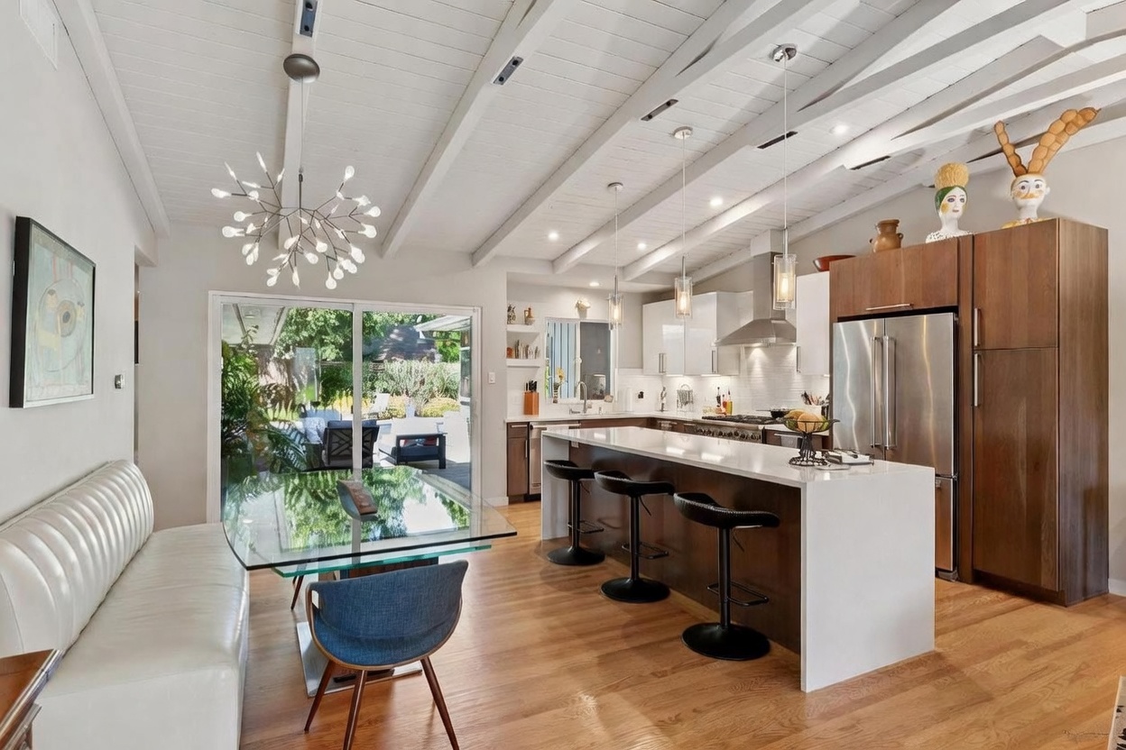 This bright, open-concept kitchen and dining area features a modern aesthetic with white vaulted ceilings, exposed beams, and warm hardwood flooring. A large central island with quartz countertops and bar seating serves as the focal point, complemented by sleek white cabinetry and stainless steel appliances. The space flows seamlessly into a dining area with a glass-topped table and a sliding glass door that provides a view of the outdoor patio.
