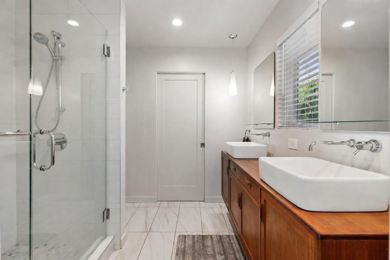 This modern primary bathroom features a sleek glass-enclosed walk-in shower with a rainfall showerhead and a double vanity with vessel sinks atop a warm-toned wooden cabinet. The space is brightened by recessed lighting and a pendant light, while white marble-style floor tiles and neutral walls create a clean, contemporary aesthetic. A large window with blinds allows natural light to filter in, enhancing the airy and sophisticated feel of the room.