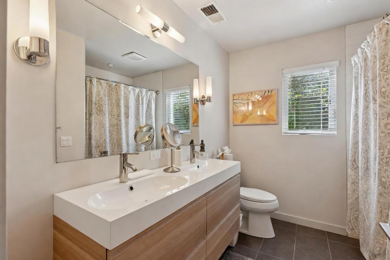 This modern primary bathroom features a sleek, double-sink vanity with light wood cabinetry and a clean white countertop. A large, wall-to-wall mirror reflects the space, while contemporary light fixtures and a neutral color palette create a bright and airy atmosphere. The room includes a tiled shower area with a patterned curtain and dark floor tiles, offering a sophisticated and functional design.
