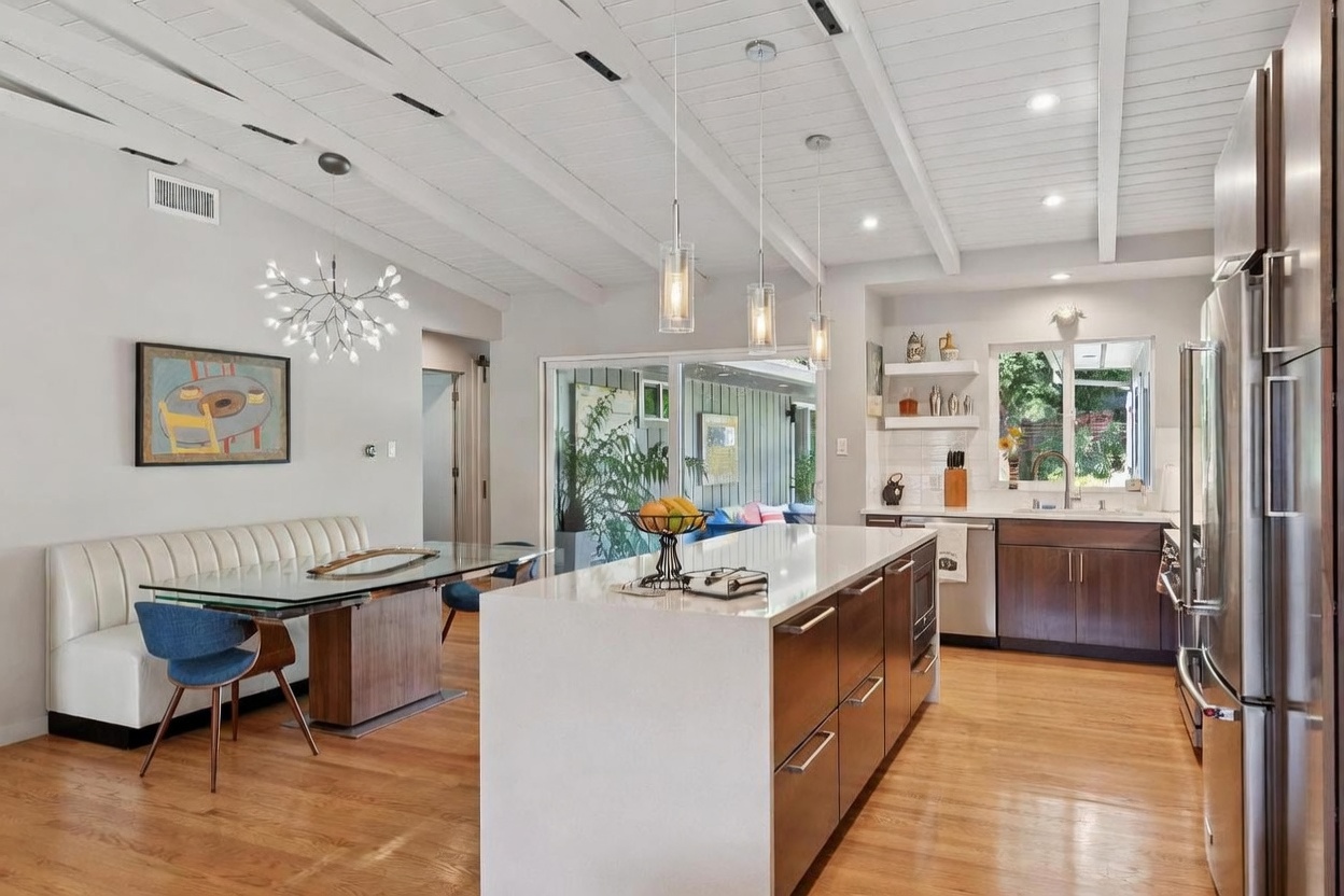 This bright, open-concept kitchen features a large central island with a white quartz countertop and dark wood cabinetry, complemented by matching wood flooring. A dining area is seamlessly integrated to the left, showcasing a modern glass-topped table, a white banquette, and stylish blue chairs under a unique sputnik-style chandelier. The space is illuminated by natural light from large sliding glass doors and a window above the sink, creating an airy and inviting atmosphere.