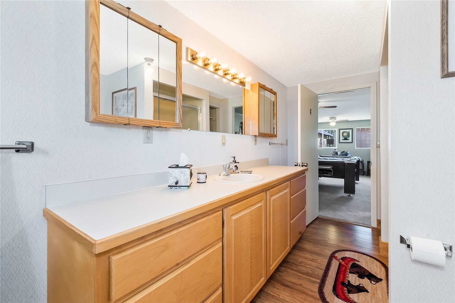 This bathroom features a light wood vanity with a solid-surface countertop and a dual-mirror medicine cabinet setup. The space is illuminated by a multi-bulb vanity light fixture above the mirror and includes a toilet paper holder and a towel bar. A doorway leads into an adjacent room, providing a view of a pool table, while a patterned rug sits on the wood-look flooring.