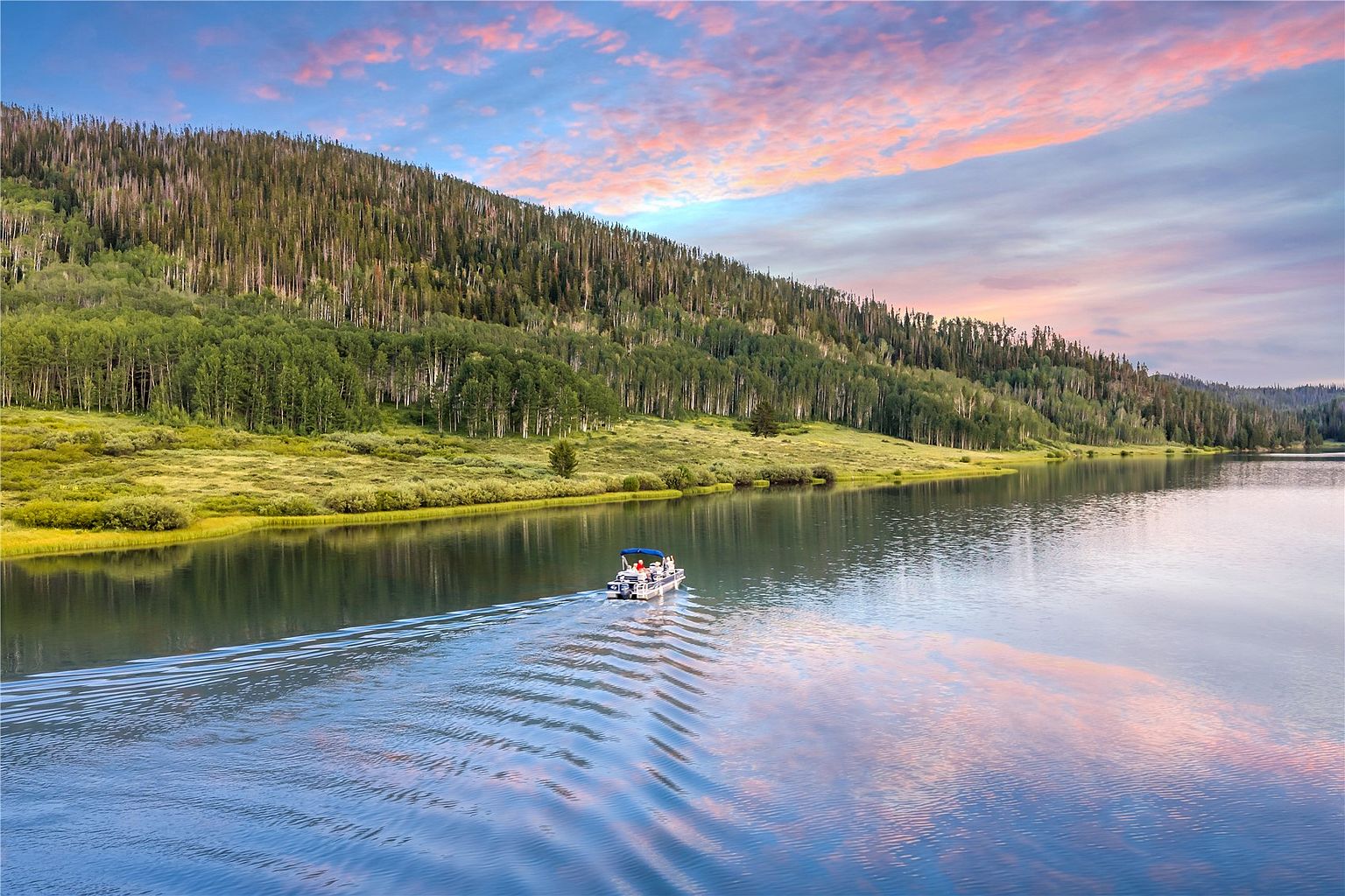 This aerial perspective captures a serene lake scene featuring a pontoon boat cruising across calm, reflective waters. The shoreline is lined with lush green vegetation and a dense forest of pine and aspen trees, set against a vibrant, colorful sunset sky. The composition emphasizes the peaceful, recreational lifestyle associated with the property's location.