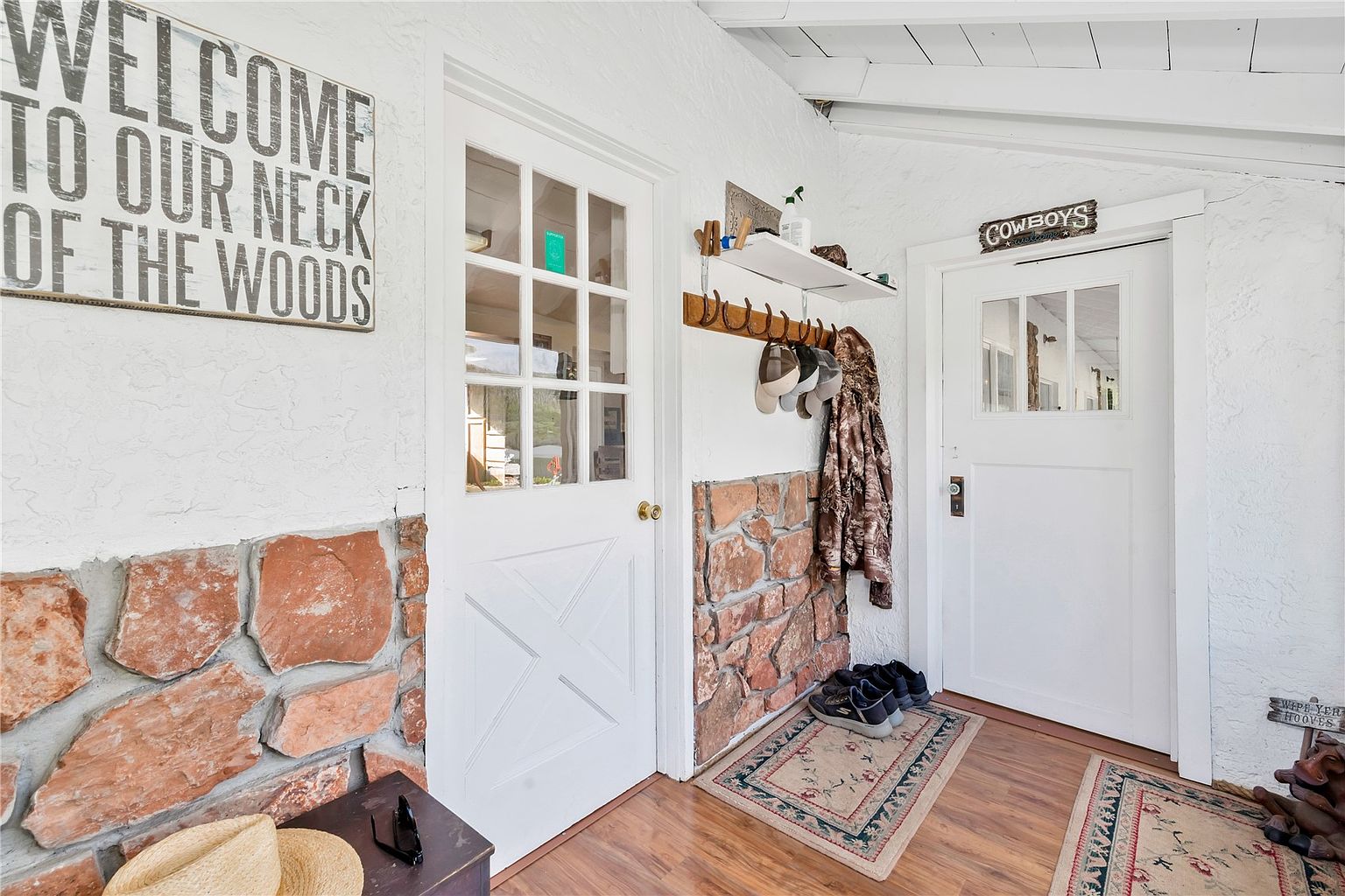 This covered entryway features a rustic, farmhouse-inspired aesthetic with white-painted textured walls and a stone wainscoting detail. The space includes two white doors with glass panes, a wooden coat rack with hooks, and a small shelf, all set against wood-look flooring with patterned area rugs. A large sign reading 'Welcome to our neck of the woods' and a 'Cowboys' plaque add a personalized, welcoming touch to the transition area.