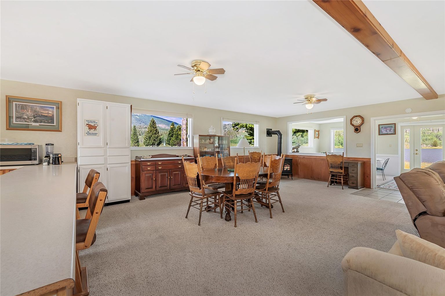 This open-concept dining area features a central wooden table surrounded by matching chairs, positioned on a neutral carpeted floor. To the left, a breakfast bar with stools connects to a kitchen area, while a wooden hutch and a desk area are placed against the walls. Large windows provide natural light and scenic views of the surrounding landscape, creating a bright and inviting atmosphere.