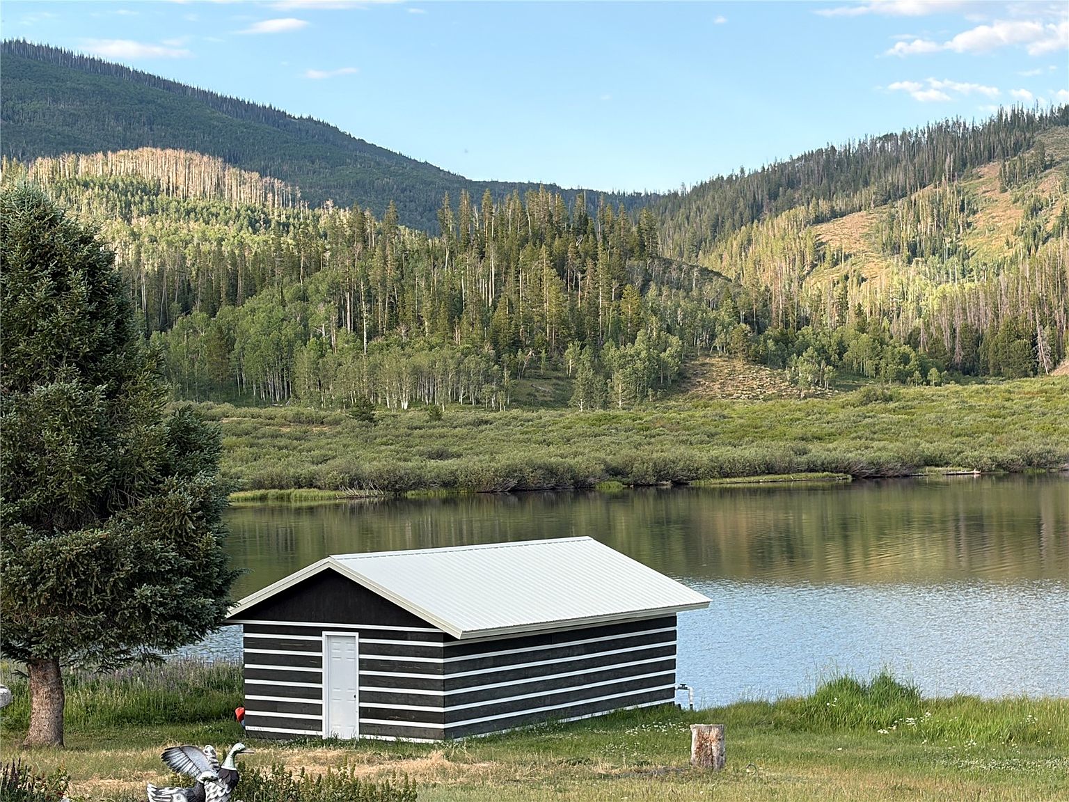 This side view captures a small, dark-sided cabin with a white metal roof situated directly on the edge of a serene lake. The structure features horizontal white trim accents and a single white door, set against a backdrop of lush green hills and a dense forest. The perspective is wide and cinematic, emphasizing the tranquil, secluded nature of the property and its immediate waterfront access.