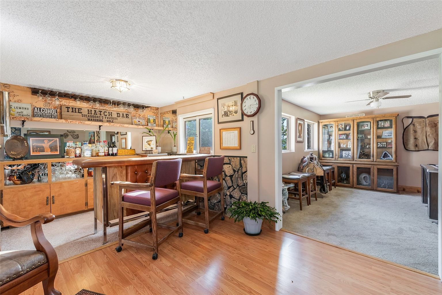 This basement space features a dedicated home bar area with a wooden counter, two bar stools, and a mirrored back wall decorated with various signs. Adjacent to the bar, a transition leads into a carpeted lounge area furnished with a large wooden display cabinet and western-themed decor, including a saddle. The room combines wood-look flooring in the bar area with neutral wall tones, creating a casual, rustic entertainment space.