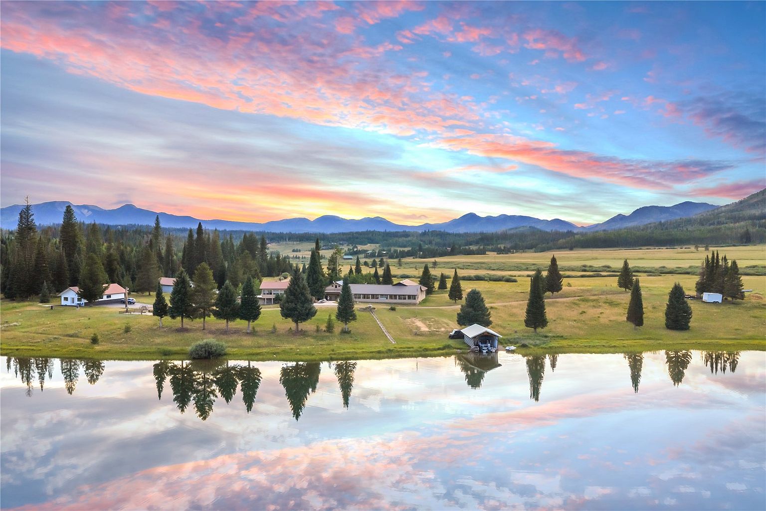 This expansive aerial view captures a serene rural property set against a backdrop of rolling mountains under a vibrant, colorful sunset sky. The landscape features a calm pond in the foreground reflecting the sky, leading to a sprawling grassy meadow dotted with mature pine trees and several residential structures. The scene conveys a peaceful, secluded atmosphere, highlighting the vast natural beauty and open space of the estate.