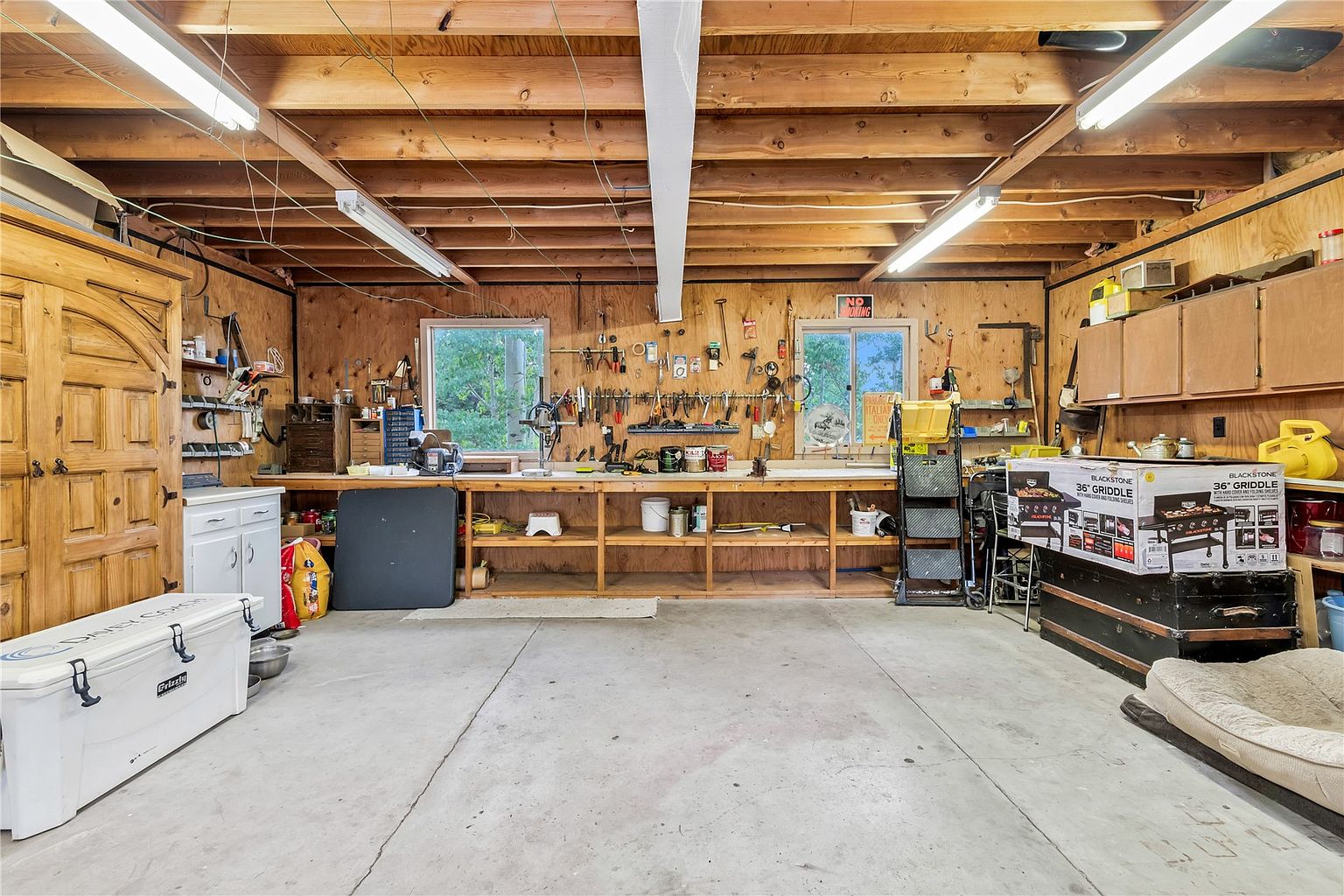 This spacious, rustic-style garage features exposed wooden ceiling beams, plywood-paneled walls, and a long, built-in workbench spanning the back wall. The space is equipped with ample overhead fluorescent lighting, a large white cooler, and a boxed Blackstone griddle, suggesting a functional workshop or hobby area. The concrete floor and open-frame construction provide a practical, utilitarian aesthetic suitable for storage or projects.