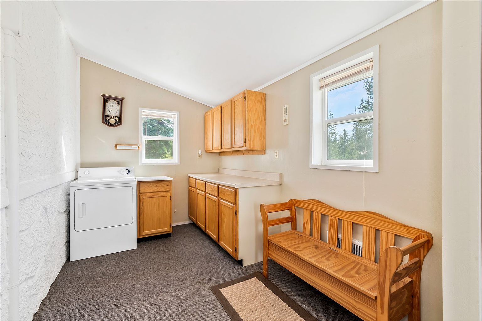 This functional laundry room features light wood cabinetry providing ample storage space above and below a laminate countertop. A white dryer is positioned to the left, while a wooden bench sits on the right side of the room, creating a convenient seating area. The space is illuminated by natural light from two windows and has a neutral, practical aesthetic.
