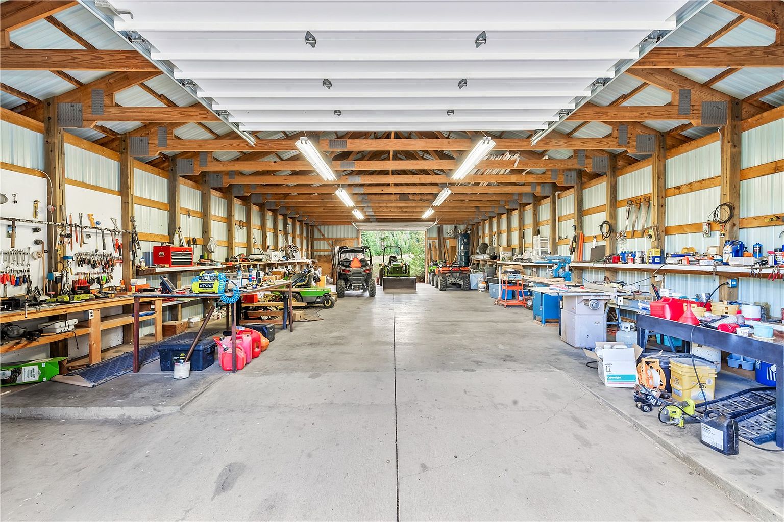 This spacious, detached workshop or garage features an open-concept design with exposed wooden rafters and metal wall paneling. The concrete floor provides ample room for vehicle storage, including ATVs and a tractor, while extensive workbench areas line both sides of the structure, equipped with various tools and storage solutions. The perspective is a wide-angle, centered view looking down the length of the building toward the open rear bay door.