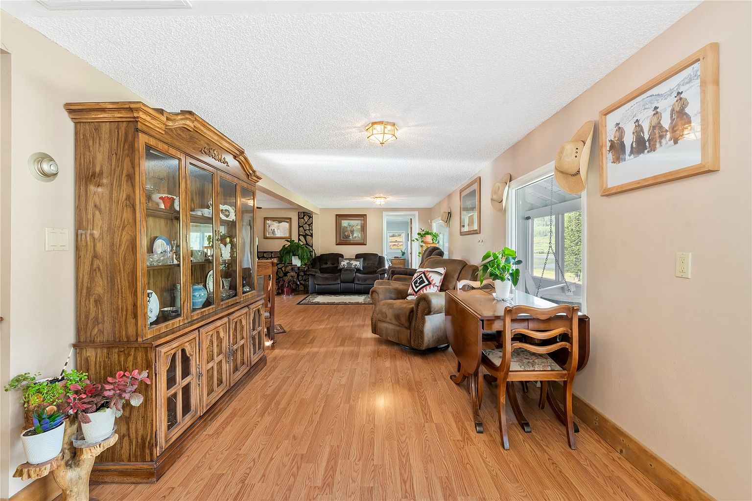 This open-concept living area features warm wood-look flooring and a traditional, cozy aesthetic. A large, ornate wooden hutch stands on the left, while a dining set with a drop-leaf table sits near a window adorned with cowboy hats. In the background, a comfortable seating area with plush brown furniture creates a welcoming space for relaxation.