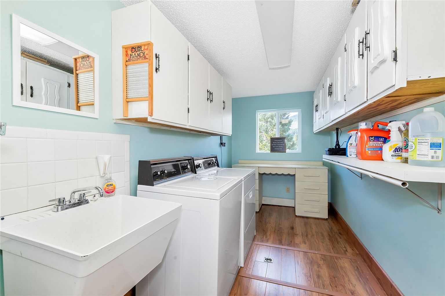 This functional laundry room features a white utility sink, a side-by-side washer and dryer set, and ample storage provided by white upper cabinets. A built-in desk area sits beneath a window at the far end of the room, while wood-look flooring and light blue walls create a bright, practical workspace. The perspective is a wide-angle shot from the doorway, showcasing the efficient layout and organization of the utility area.