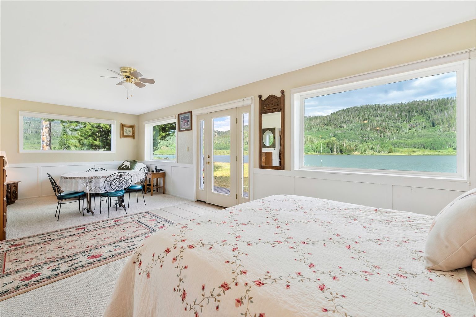 This bright bedroom features a large bed with a floral patterned quilt, positioned to take advantage of the expansive lake and mountain views through a wide window. A small dining area with a round table and chairs sits in the corner, creating a versatile living space. The room is finished with neutral walls, white wainscoting, and a glass door that provides direct access to the outdoors.