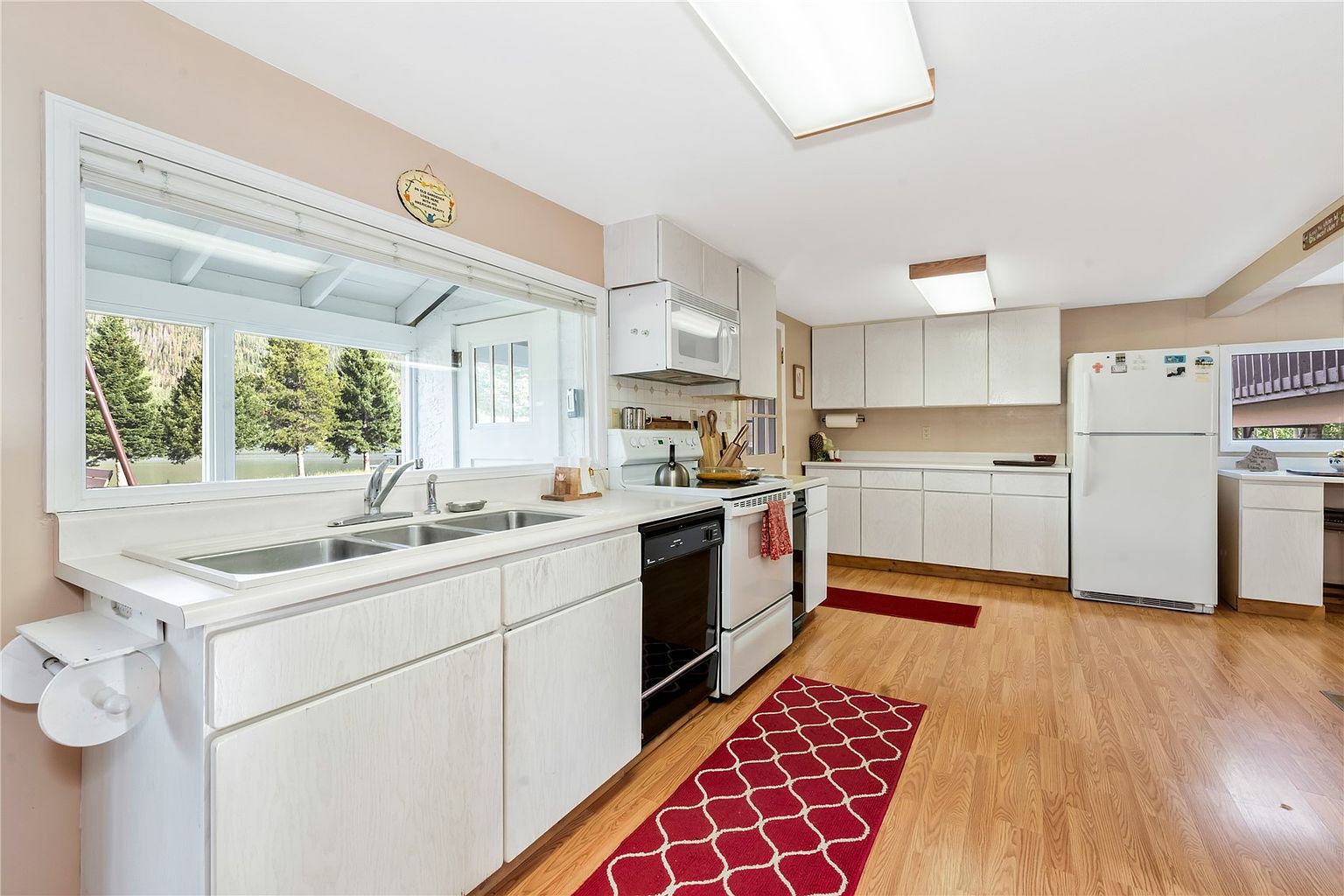 This kitchen features white cabinetry, light wood-toned flooring, and a large window above the sink that provides a scenic view of the outdoors. The space is equipped with a white stove, a black dishwasher, and a white refrigerator, complemented by red patterned runner rugs. The perspective is a wide-angle shot from the kitchen entrance, capturing the functional layout and the bright, airy atmosphere.