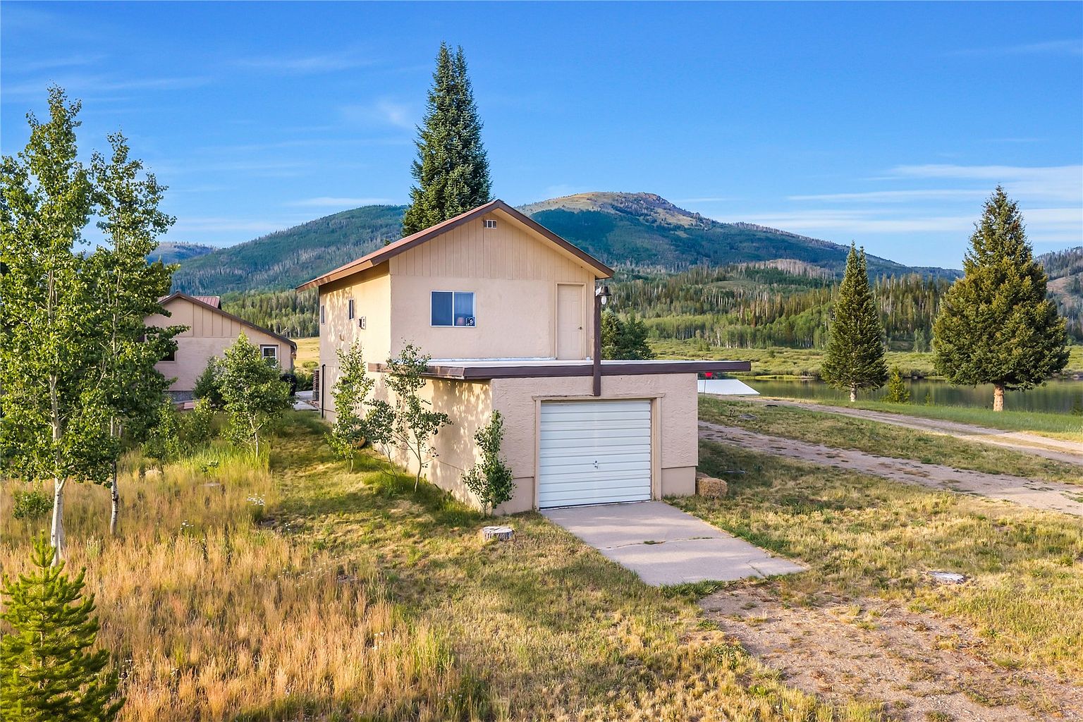 This side-view perspective captures a two-story structure featuring a ground-level garage with a white roll-up door and an upper-level living area with a single window and a side door. The building is set against a scenic backdrop of rolling mountains, evergreen trees, and a nearby lake, emphasizing a peaceful, rural setting. The exterior is finished in a neutral tone, and the property is surrounded by natural, grassy terrain.