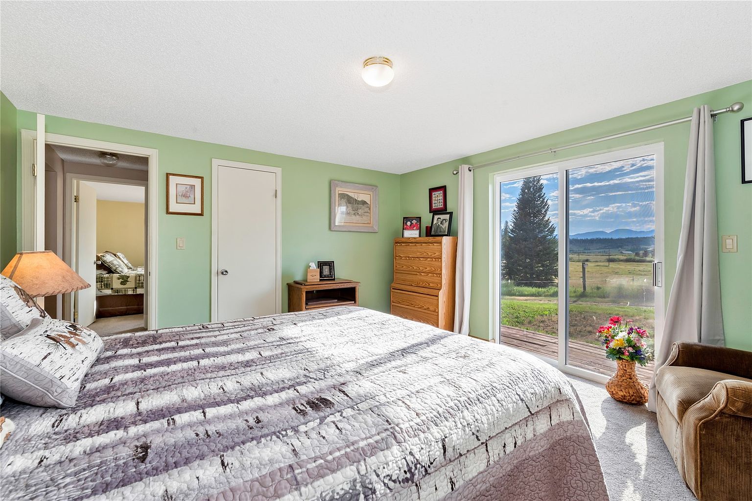 This primary bedroom features light green walls and a large bed with a patterned quilt, creating a cozy and inviting atmosphere. A sliding glass door provides access to an outdoor deck and offers expansive views of the surrounding landscape. The room is furnished with a wooden dresser and a comfortable armchair, with an open doorway leading to an adjacent bedroom.
