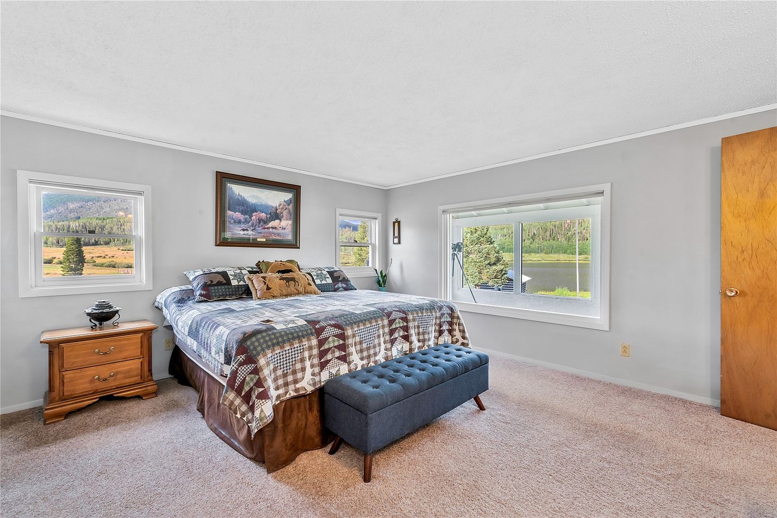 This primary bedroom features a rustic, cabin-inspired aesthetic with a large bed adorned in a patchwork quilt and bear-themed accent pillows. The room is illuminated by natural light from multiple windows that offer scenic views of the surrounding landscape and lake. A wooden nightstand and a tufted blue bench at the foot of the bed complete the cozy, comfortable atmosphere.