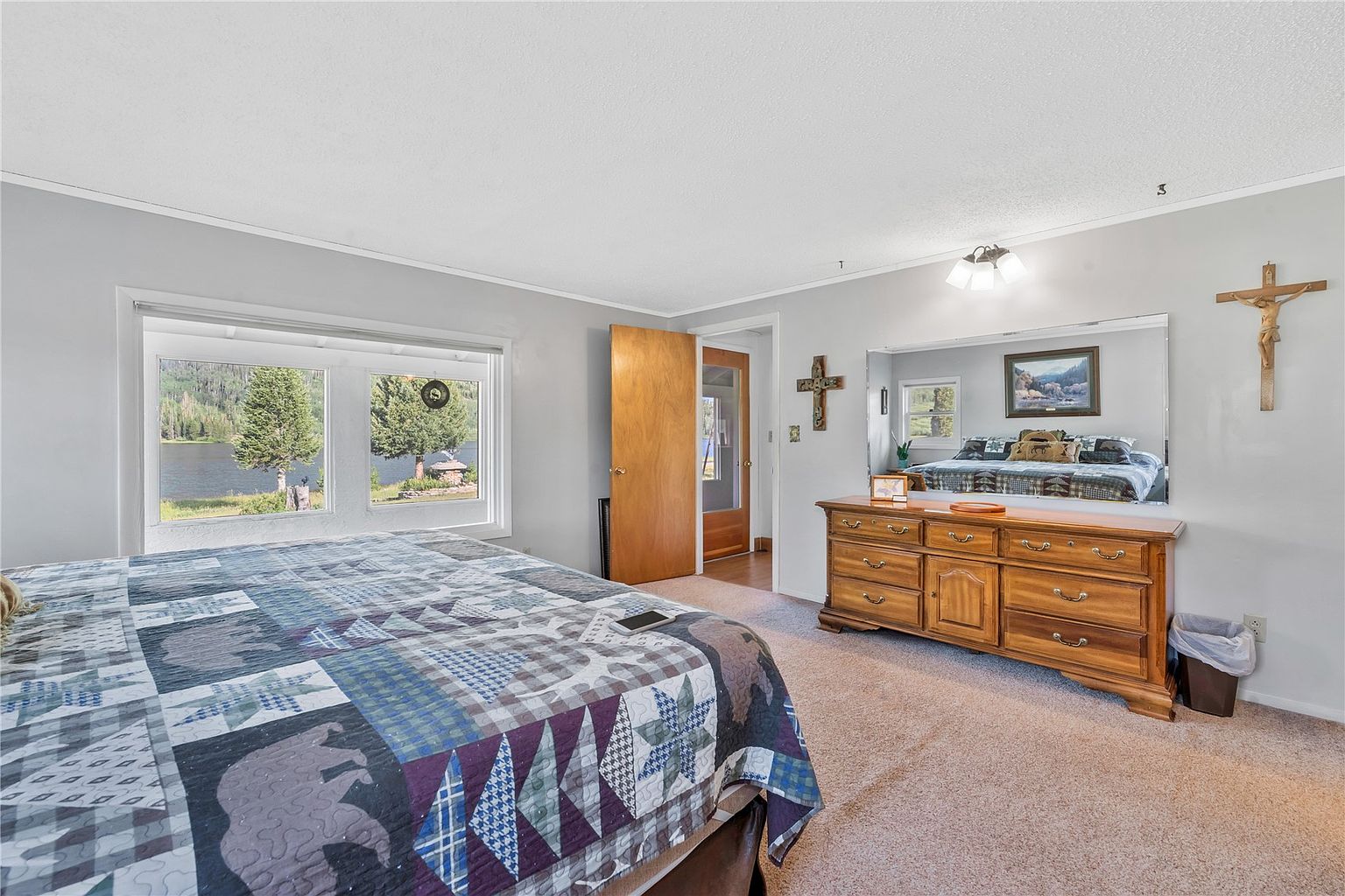 This bedroom features a rustic aesthetic with a patterned quilt on the bed and a large wooden dresser topped with a mirror. A wide window provides a scenic view of a lake and surrounding trees, while religious wall decor adds a personal touch. The room is carpeted and includes a doorway leading to an adjacent space, creating a cozy and traditional atmosphere.