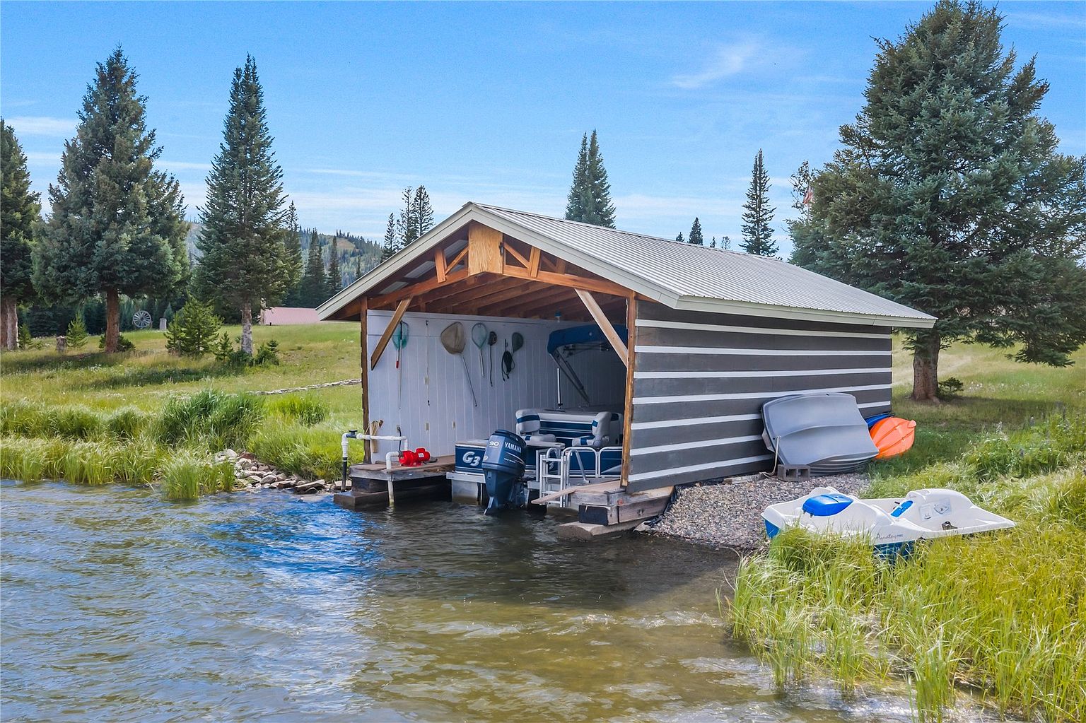 This image captures a rustic boat house situated directly on the edge of a calm lake, surrounded by lush green grass and tall pine trees. The structure features a simple gabled roof, horizontal siding, and an open front that houses a pontoon boat with a Yamaha motor. A small pedal boat rests on the rocky shoreline in the foreground, emphasizing the property's direct water access and recreational appeal.