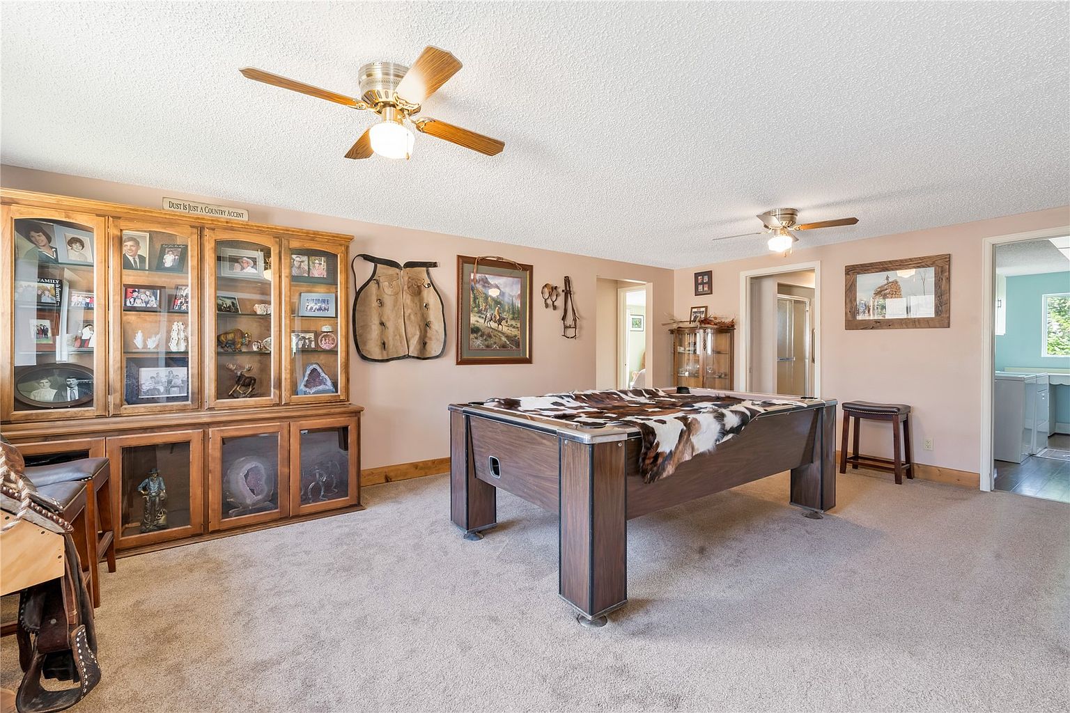 This spacious recreation room features a large pool table draped with a cowhide cover as the central focal point. The room is decorated with a western-inspired theme, including a large wooden display cabinet filled with collectibles, wall-mounted chaps, and framed artwork. The space is illuminated by two ceiling fans and features neutral carpeting, creating a comfortable and inviting area for entertainment.