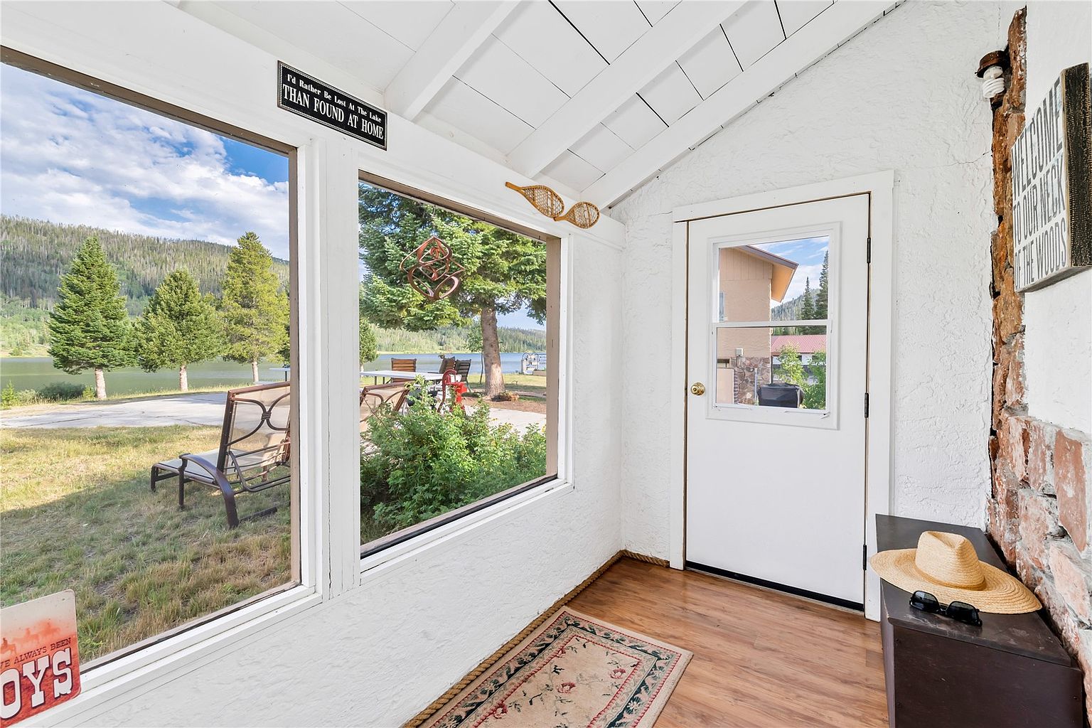 This bright sunroom features white-painted walls and a vaulted ceiling, creating an airy and inviting atmosphere. Large windows provide expansive views of the lake and surrounding forest, while a white door leads to the exterior. A rustic stone-clad wall section, a straw hat on a small bench, and decorative wall hangings contribute to a cozy, cabin-like aesthetic.