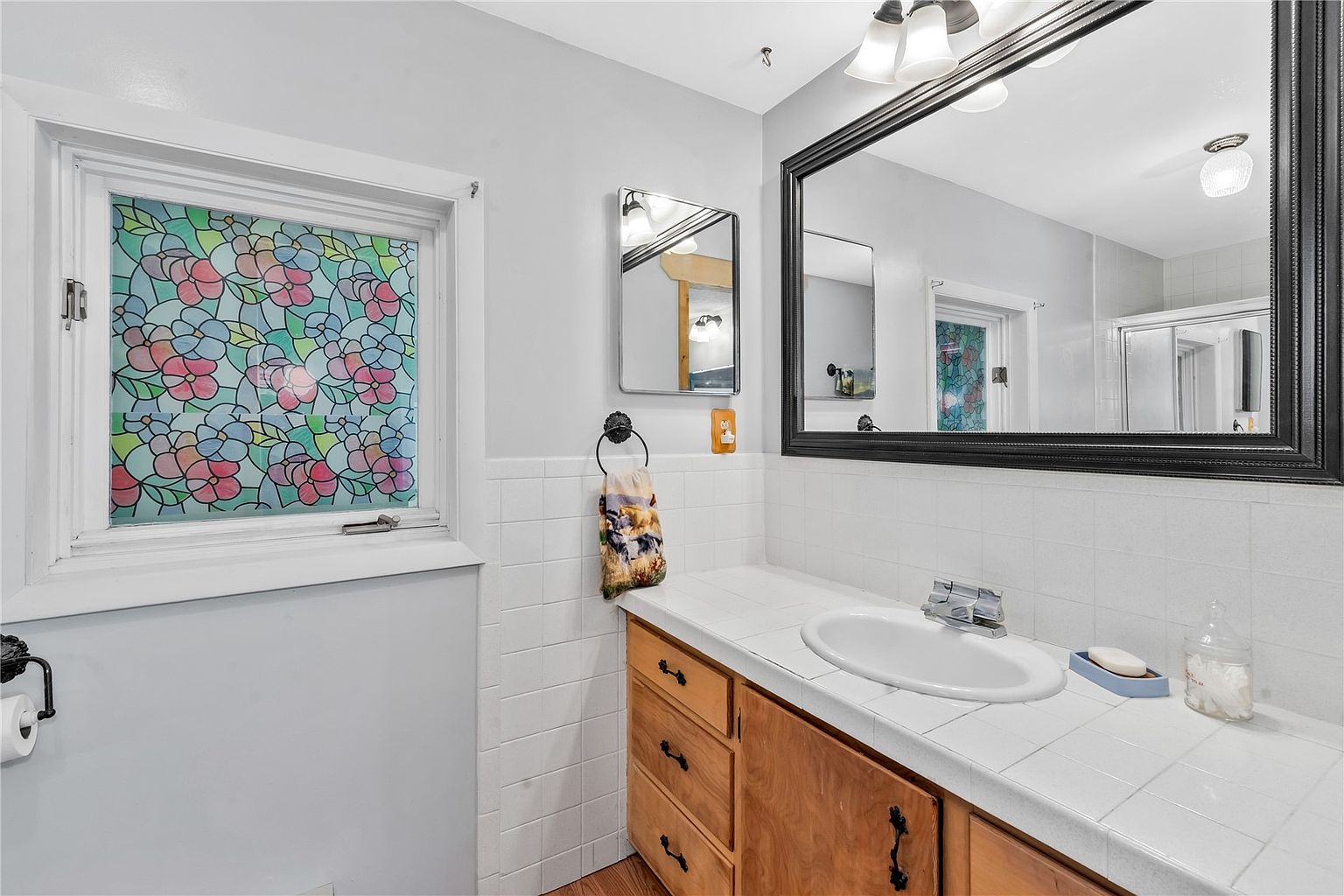 This bathroom features a vanity with a tiled countertop and light wood cabinetry, paired with a large mirror framed in black. A window with a colorful floral-patterned privacy film adds a unique decorative touch to the space. The walls are painted a soft gray, complemented by white wall tiles surrounding the vanity area.