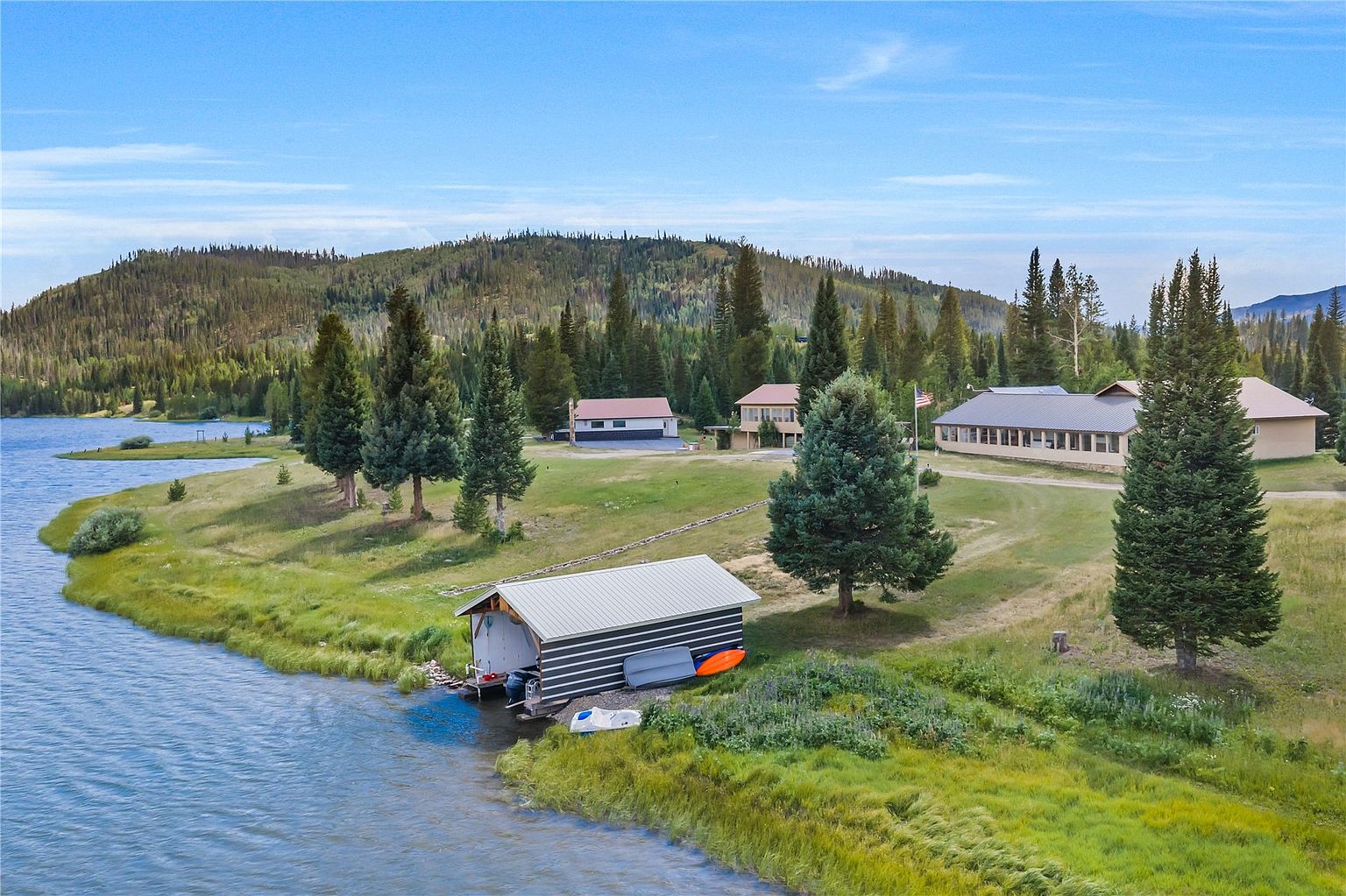This aerial perspective captures a serene lakeside property featuring a boat house at the water's edge and several residential buildings nestled among mature pine trees. The expansive grassy landscape slopes gently toward the lake, framed by a backdrop of rolling, forested hills under a clear blue sky. The scene conveys a peaceful, secluded retreat atmosphere, ideal for outdoor recreation and waterfront living.