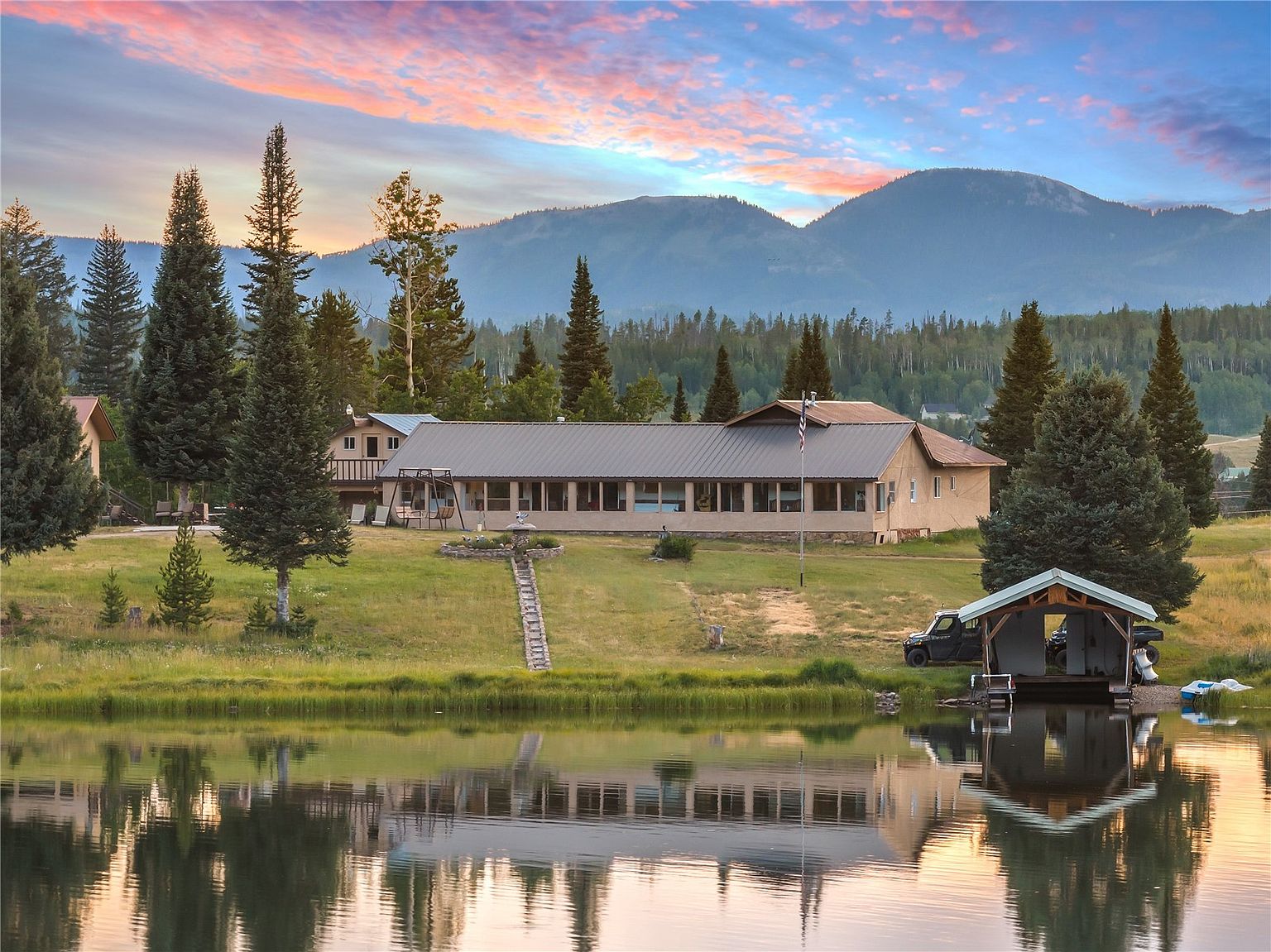 This scenic rear view captures a sprawling single-story home situated along the edge of a calm, reflective lake. The property features a long, horizontal structure with a metal roof, large windows, and a stone-lined pathway leading from the water's edge up to the house. Surrounded by lush pine trees and rolling hills under a vibrant sunset sky, the setting offers a peaceful, rustic retreat atmosphere.