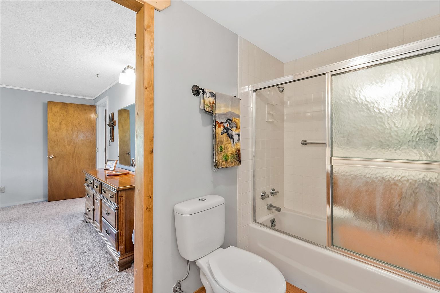 This bathroom features a white toilet and a shower-tub combination enclosed by sliding glass doors with a textured finish. The space is adjacent to a bedroom area, visible through an open doorway, which contains a wooden dresser and carpeted flooring. The neutral wall color and functional layout provide a practical and accessible design.