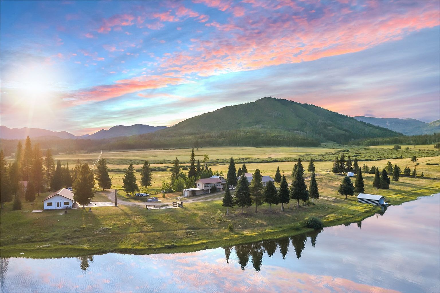 This expansive aerial view captures a serene rural property nestled against a backdrop of rolling mountains and a vibrant sunset sky. The landscape features a large, calm lake in the foreground, multiple residential structures, and a vast open meadow dotted with evergreen trees. The scene conveys a peaceful, secluded lifestyle with ample space and natural beauty.