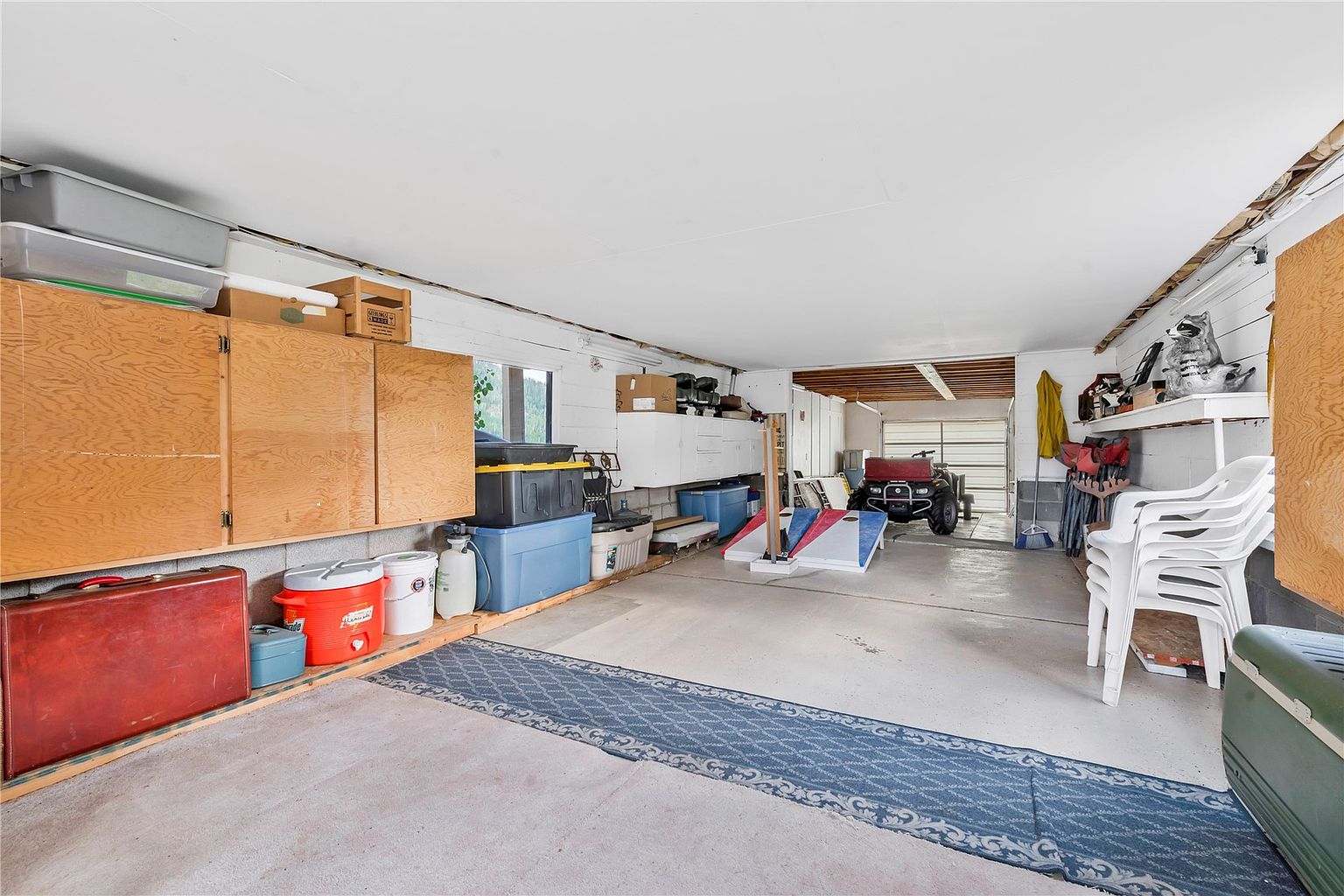 This spacious, multi-purpose garage features a concrete floor covered by a long blue patterned runner rug, providing ample storage and utility space. The area is equipped with light wood cabinetry, various storage bins, and a stack of white plastic chairs, with a view leading toward an open bay containing an ATV. The perspective is a wide-angle shot from the interior looking toward the garage door, highlighting the functional layout and depth of the storage area.