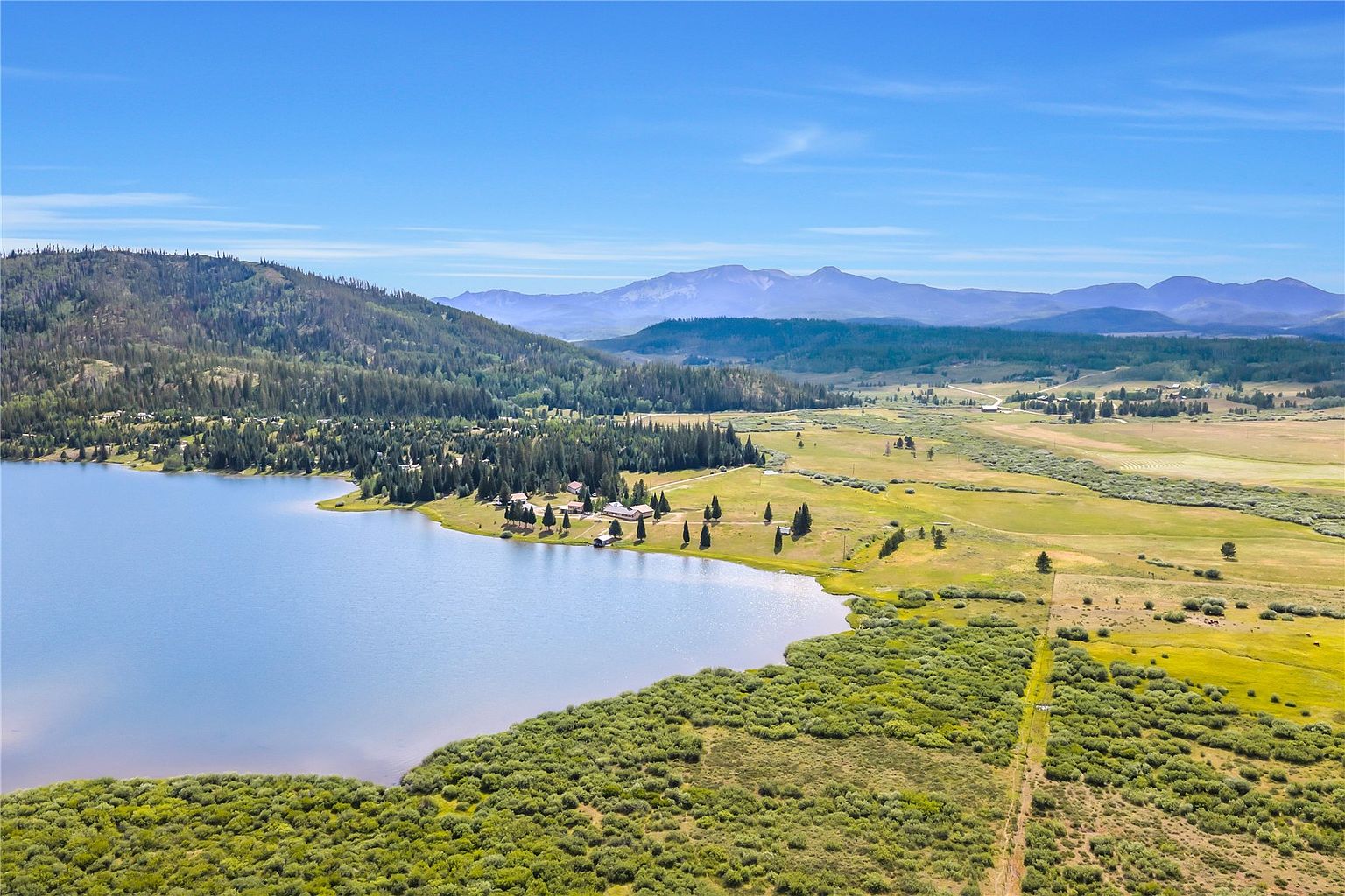 This expansive aerial view captures a serene lakeside property nestled within a vast, mountainous landscape. The scene features a large, calm lake bordered by dense evergreen forests, open grassy meadows, and a small cluster of buildings situated along the shoreline. The composition emphasizes the property's remote, natural beauty and its integration into the surrounding wilderness.