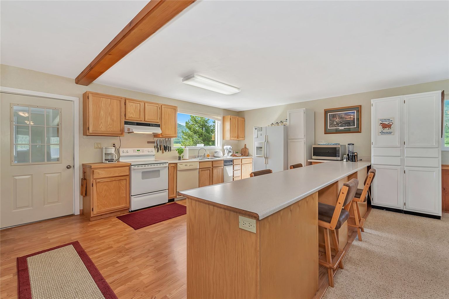This kitchen features light wood cabinetry, a white stove, and a central island with bar seating, creating a functional and open layout. A large window above the sink provides natural light and a view of the outdoors, while a wooden ceiling beam adds a rustic touch to the space. The room transitions from a hardwood floor area in the kitchen to a carpeted dining or living space, offering a comfortable and practical design.
