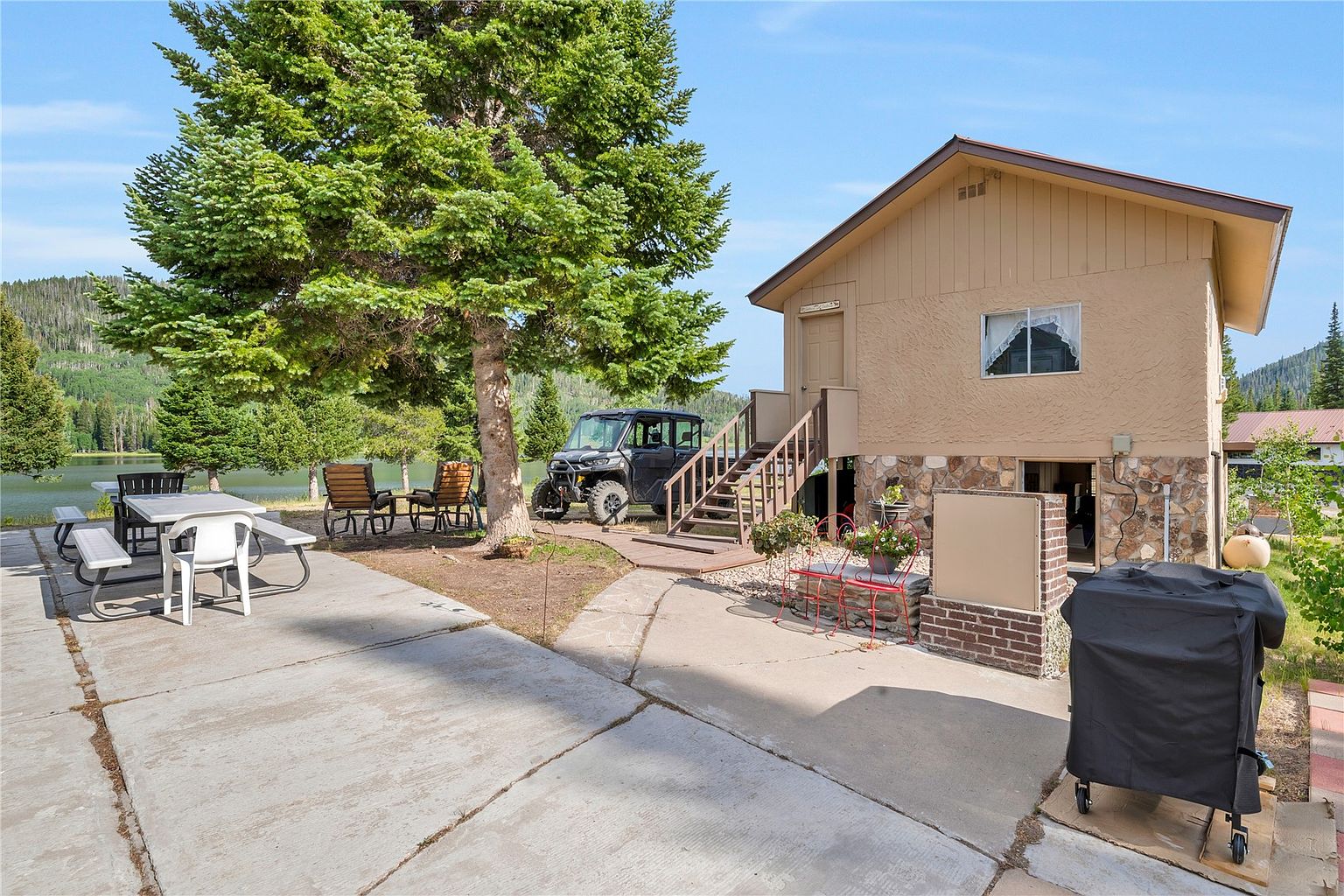 This outdoor living space features a concrete patio area equipped with a picnic table and a grill, set against a backdrop of a lake and forested mountains. A two-story cabin with a stone-clad lower level and wood-sided upper level sits adjacent to the patio, featuring an elevated wooden staircase leading to the main entrance. A utility vehicle is parked near a large, mature pine tree, creating a rustic and inviting atmosphere perfect for lakeside recreation.